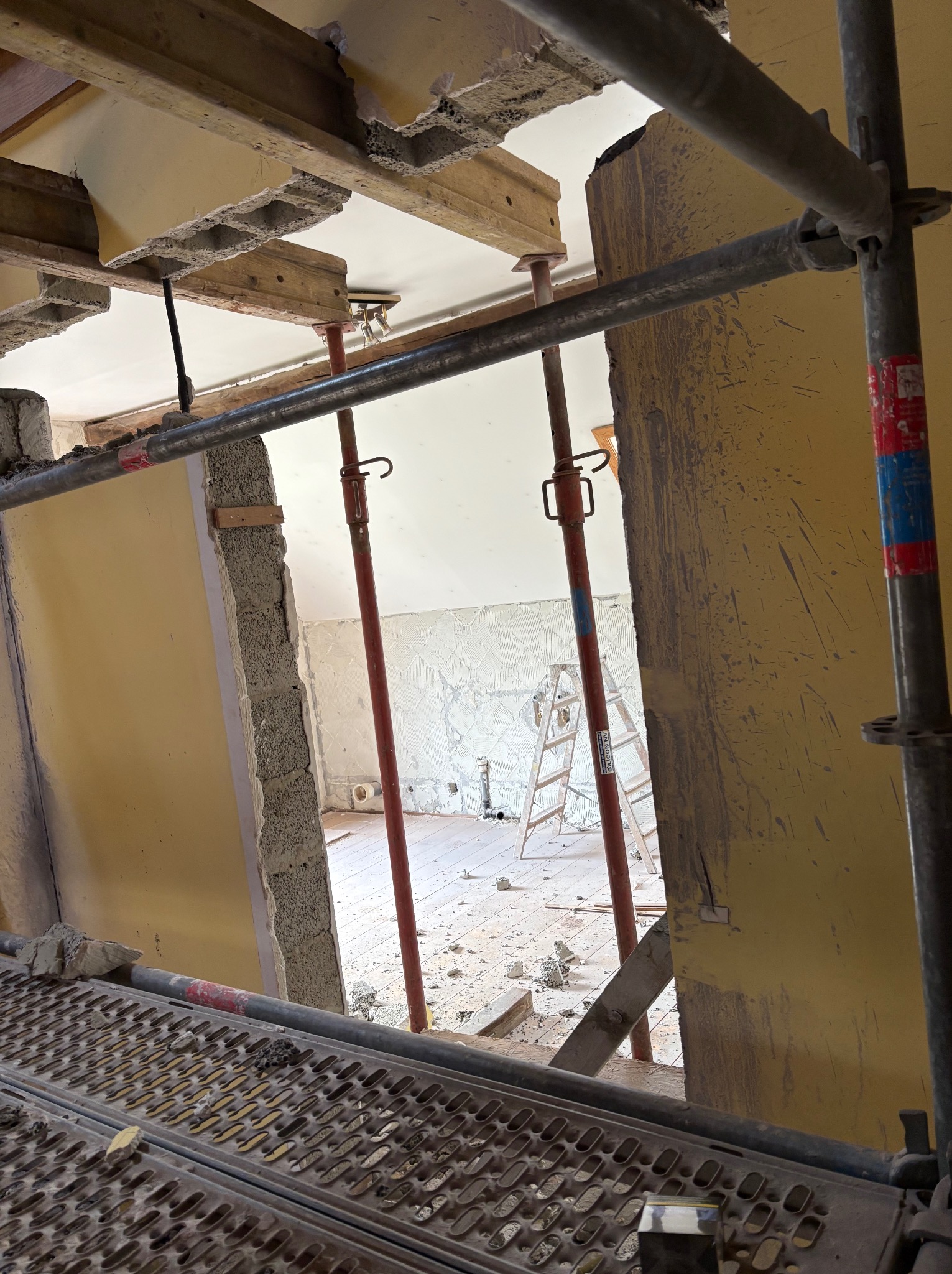 Wnętrze w trakcie demontażu: widoczne rusztowanie, podparte stropy, ściany w trakcie skuwania, gruz na podłodze, drabina. W tle pomieszczenie z częściowo położoną gładzią.