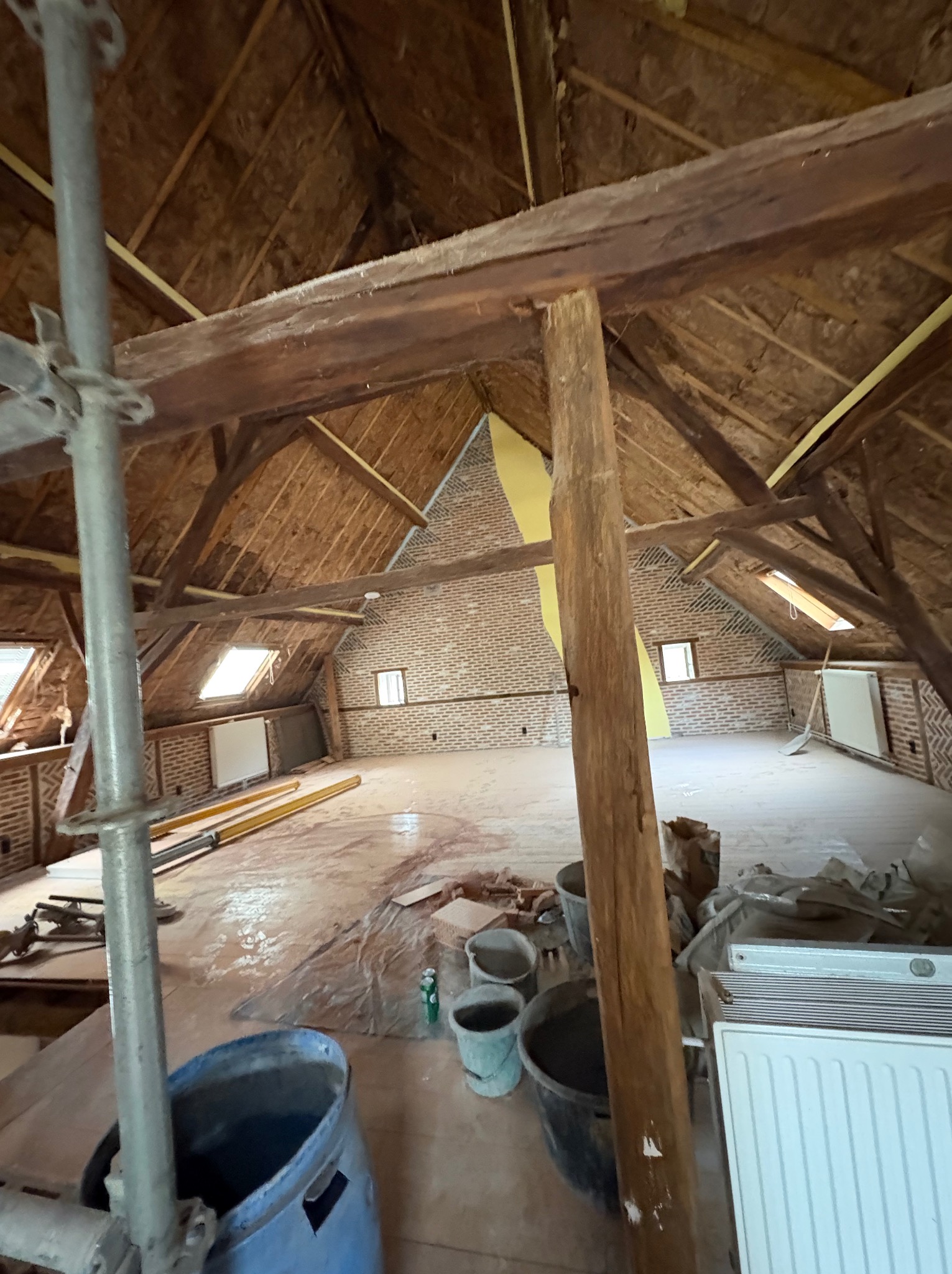 Poddasze w trakcie remontu: widoczne drewniane belki, ceglana ściana, okna dachowe, narzędzia i materiały budowlane. W tle grzejnik i przygotowane podłoże.