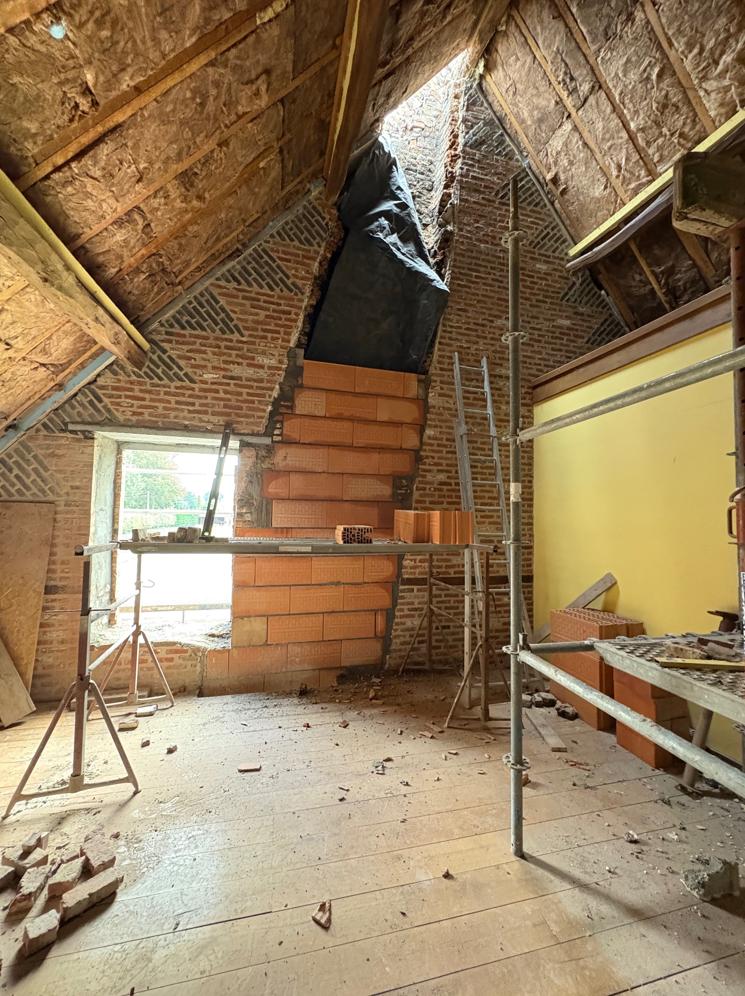 Poddasze w trakcie przebudowy: widoczne cegły, rusztowania, izolacja dachu i okno. Częściowo wykończona ściana z cegły ceramicznej.