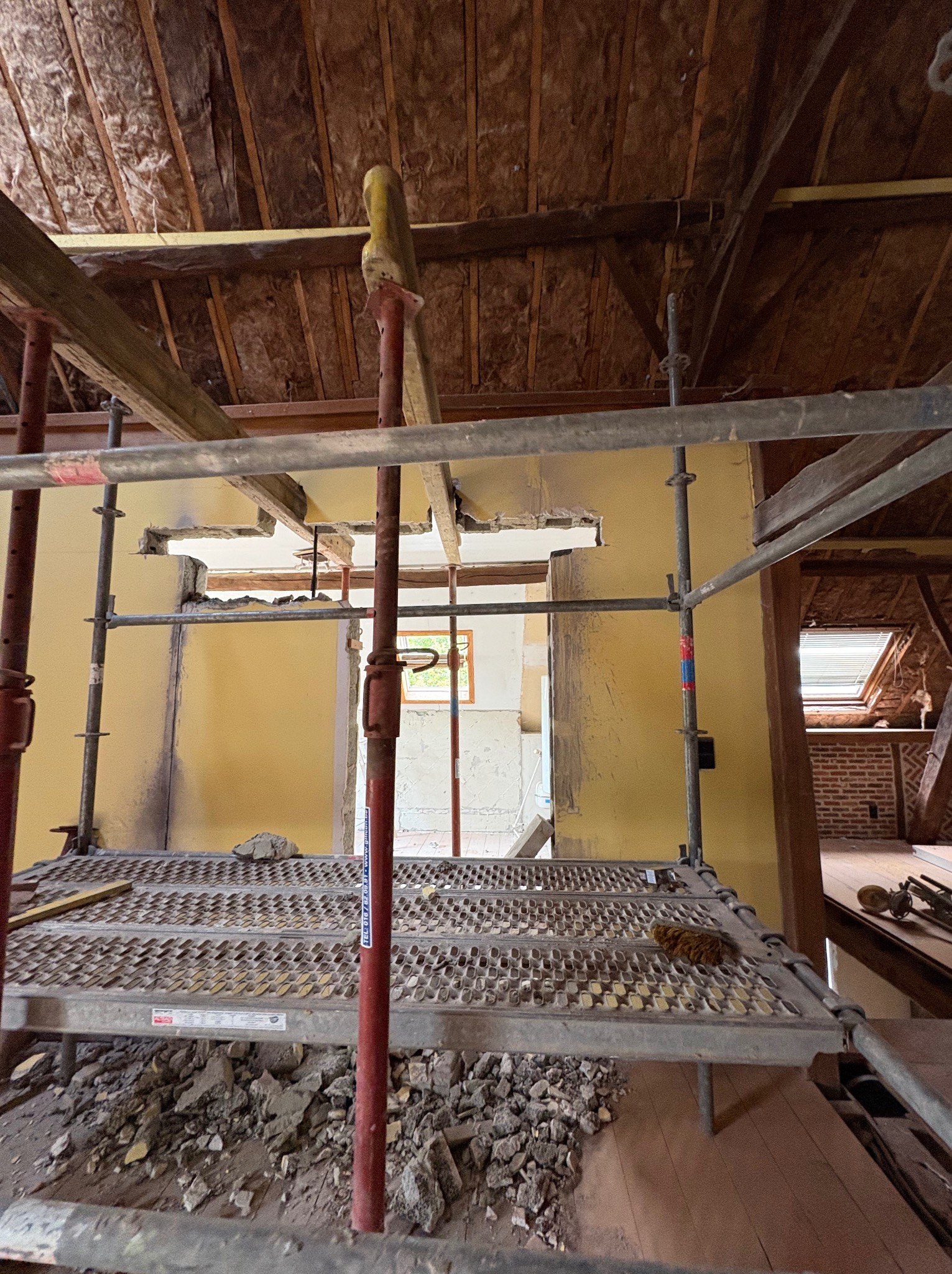 Wnętrze poddasza w trakcie przebudowy: rusztowanie, odsłonięta izolacja dachu, fragmenty ścian w kolorze żółtym. Widoczne ślady po demontażu elementów konstrukcyjnych.