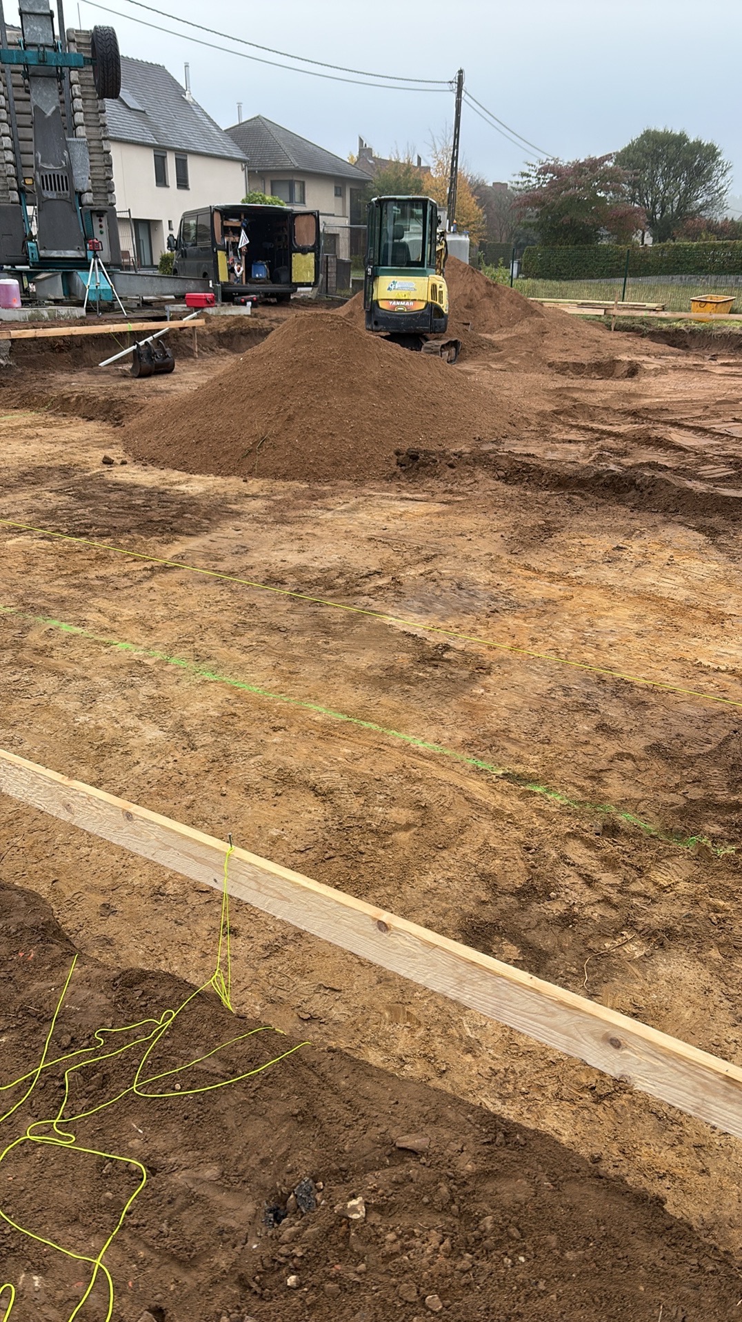 Przygotowanie terenu pod budowę: wykopane fundamenty, drewniane szalunki, mini koparka i zaparkowany bus na działce. Widok na domy w tle.