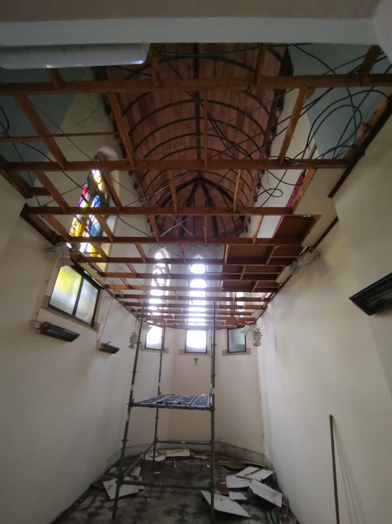 Wnętrze sakralne w trakcie prac remontowych. Widoczne rusztowanie, odsłonięta konstrukcja sufitu z elementami instalacji elektrycznej, okna witrażowe. Nierówna podłoga.