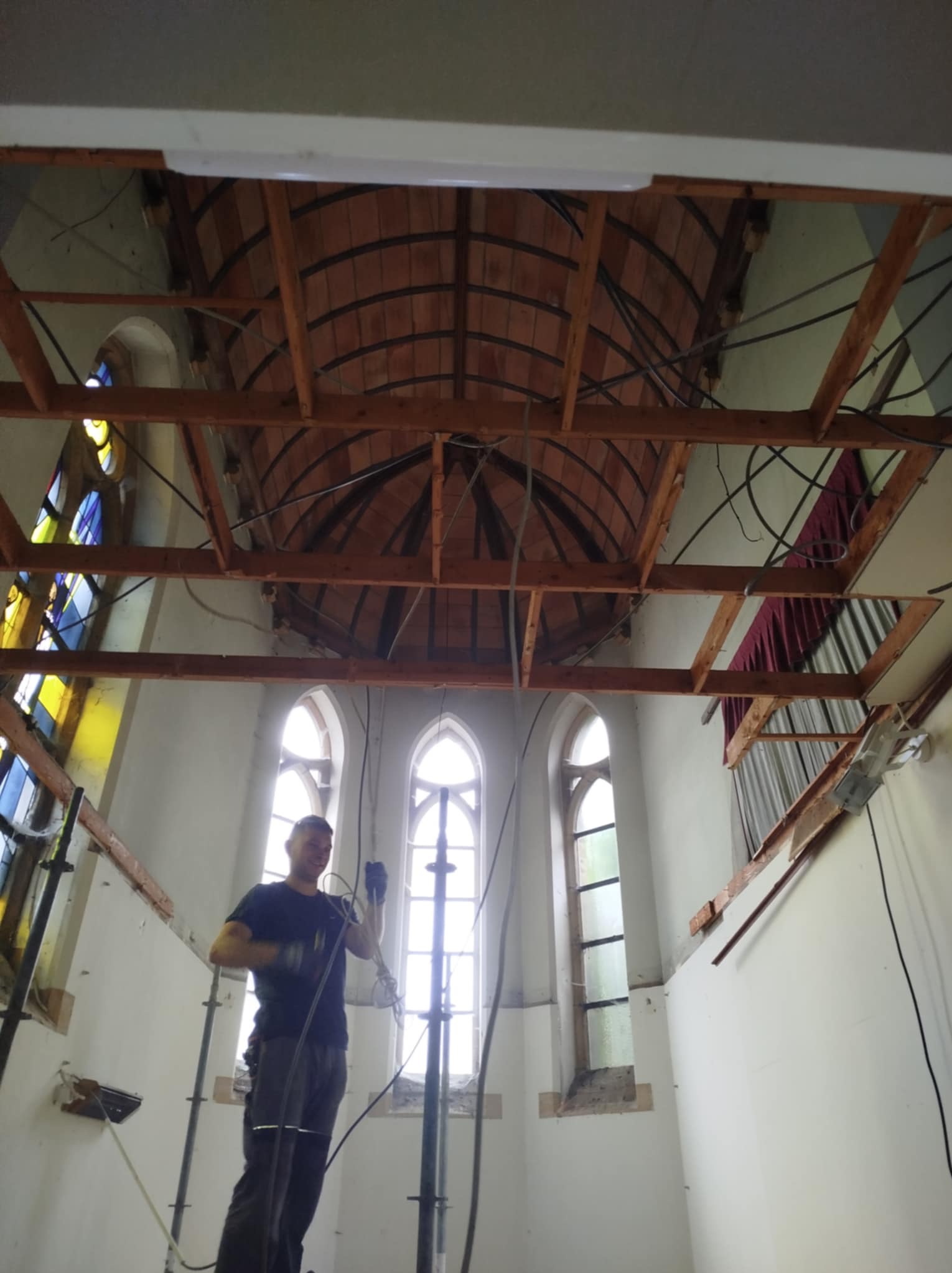 Demontaż sufitu w kościele. Widoczne drewniane elementy konstrukcyjne i fragmenty starej instalacji. Pracownik w trakcie pracy, okna witrażowe w tle.
