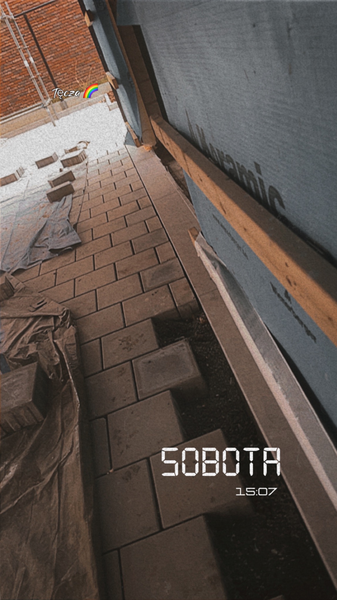 Układanie kostki brukowej na balkonie, widok z góry pod kątem. Częściowo przykryte folią, obok ściana z izolacją. W tle ceglany budynek.