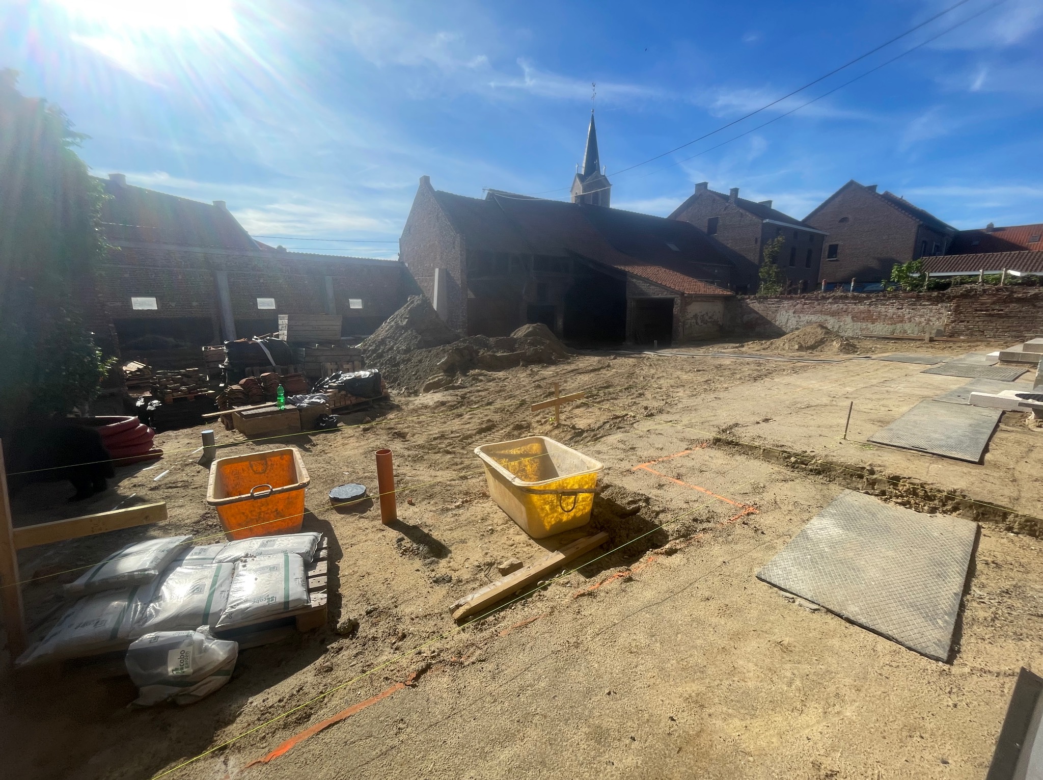 Teren budowy w słoneczny dzień: przygotowany grunt, worki z materiałem, plastikowe pojemniki i zarys fundamentów budynku z cegły w tle.