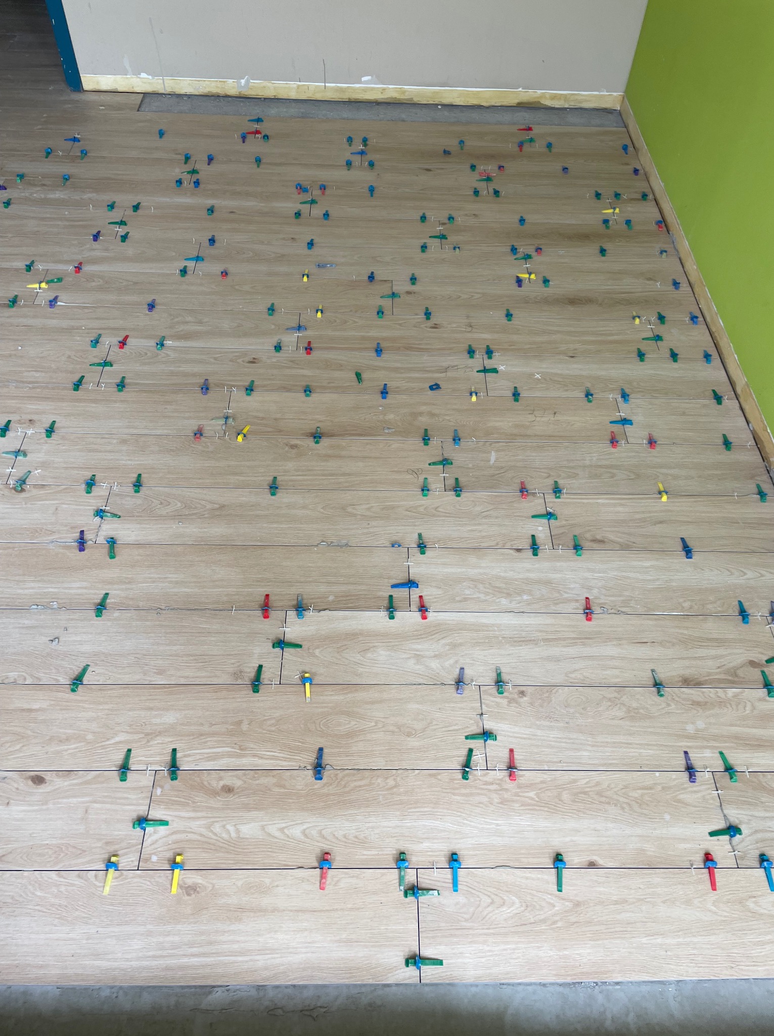 Układanie podłogi z płytek imitujących drewno, z użyciem kolorowych klipsów poziomujących. Widoczne fragmenty ścian w odcieniach beżu i zieleni.