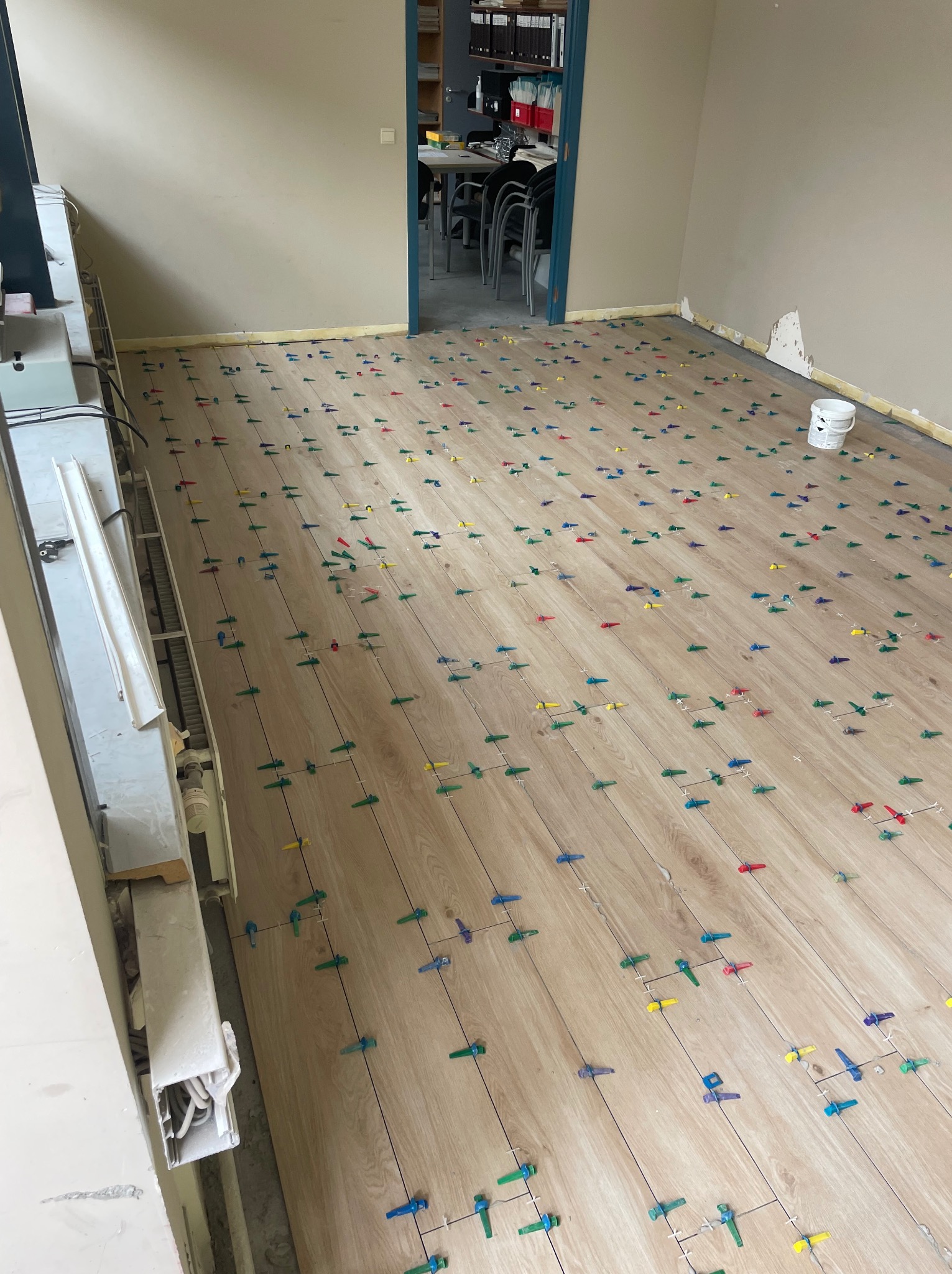 Układanie paneli podłogowych z użyciem kolorowych klipsów poziomujących w jasnym pomieszczeniu biurowym. Widoczne listwy przypodłogowe i grzejnik.