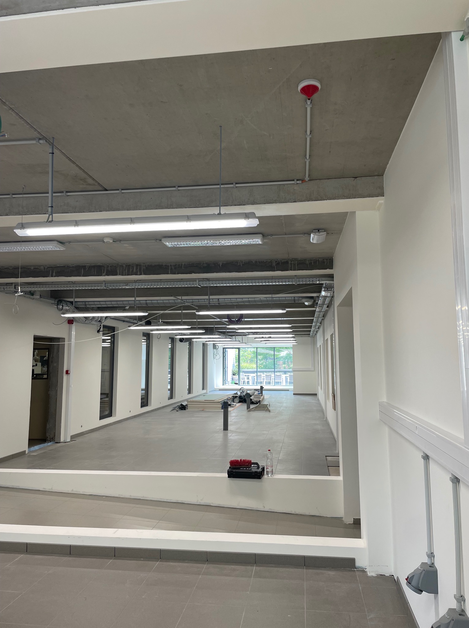 Wnętrze biura w trakcie prac wykończeniowych: szara posadzka, białe ściany, surowy betonowy sufit z instalacjami. Widoczne narzędzia i materiały budowlane w oddali.