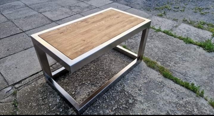 Stolik kawowy z blatem z jasnego drewna i metalową ramą, ustawiony na szarej, betonowej posadzce z widoczną zielenią. Nowoczesny design.