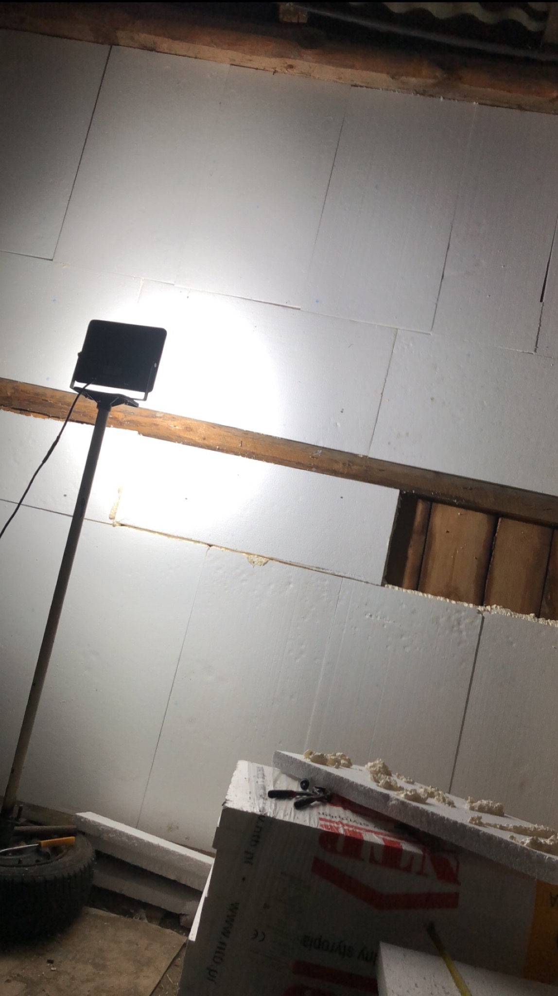 Montaż płyt styropianowych na ścianie z drewnianą belką. Widoczna lampa oświetlająca miejsce pracy i narzędzia na styropianie. Etap ocieplania pomieszczenia.