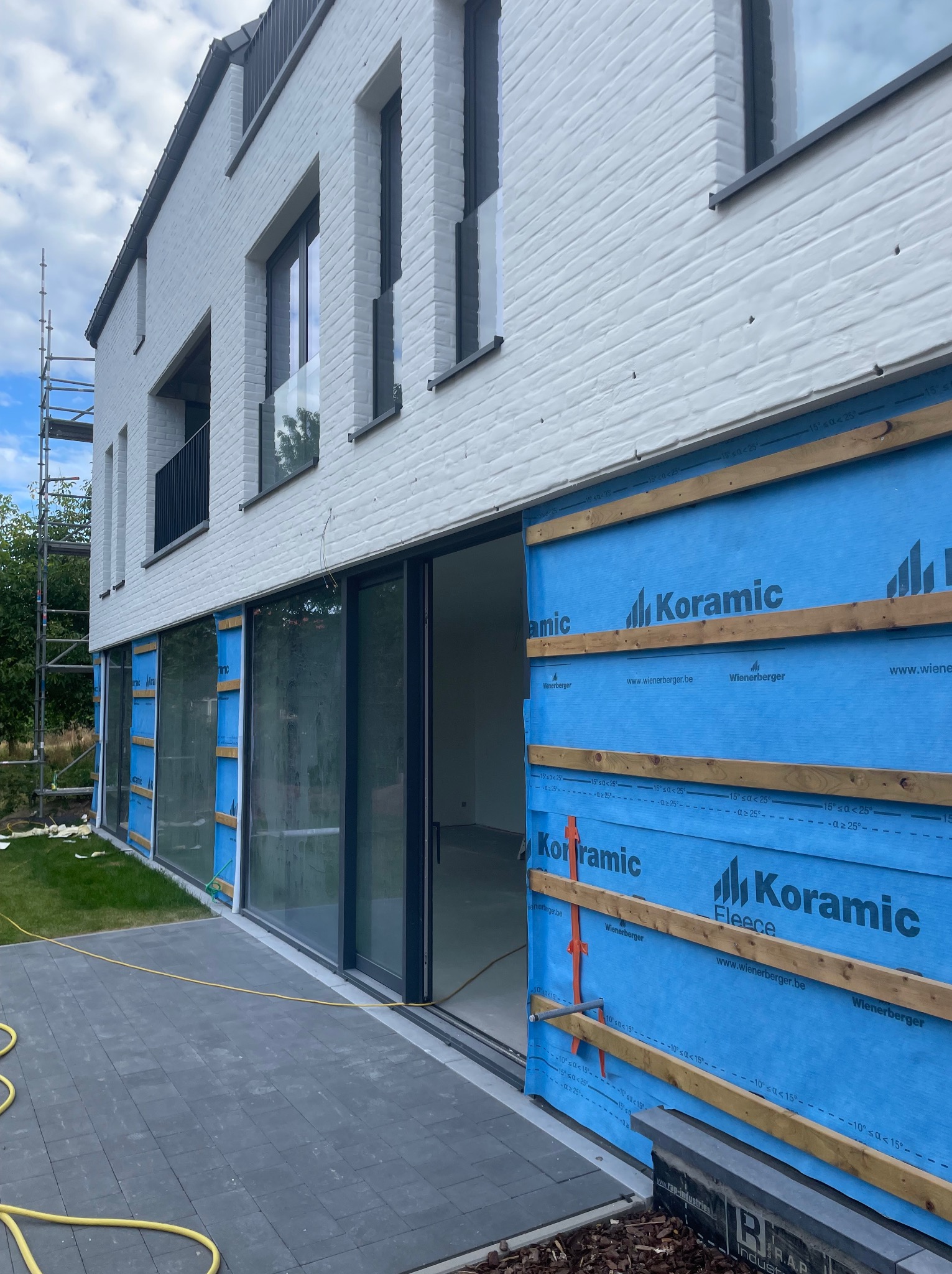 Montaż elewacji budynku z widoczną warstwą izolacyjną (niebieska membrana) i drewnianymi łatami. Nowoczesna architektura, biała cegła, duże okna przesuwne.