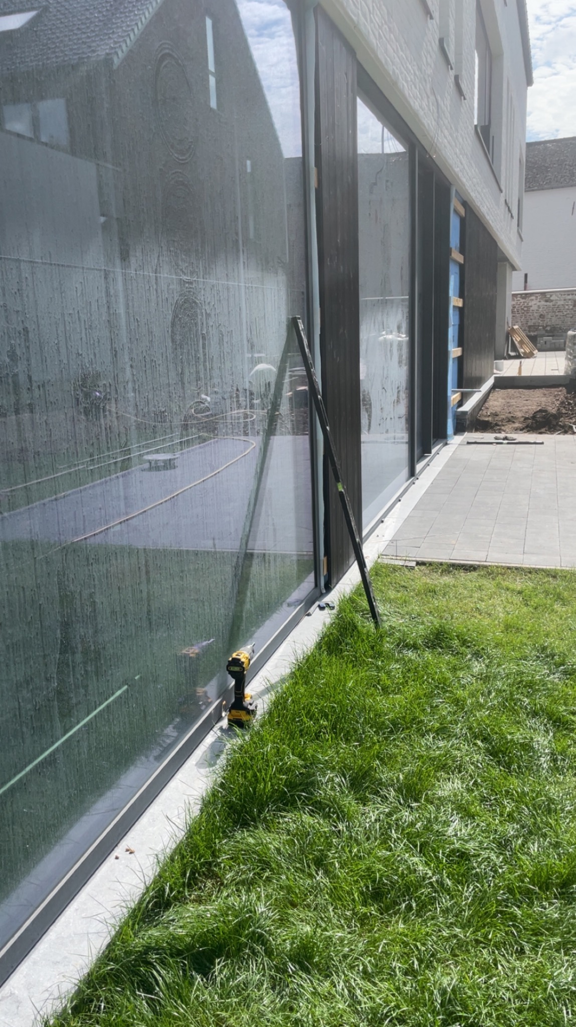 Montaż okien przesuwnych w nowoczesnym domu. Widoczny profil okienny, narzędzia i trawnik. Okna odbijają otoczenie, tworząc ciekawy efekt wizualny.