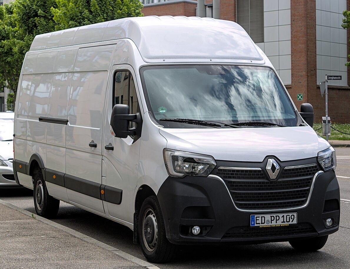 Renault Master w miejskiej scenerii, z widocznym numerem rejestracyjnym i odbiciem chmur na szybie, zaparkowany przy chodniku z zielenią w tle.