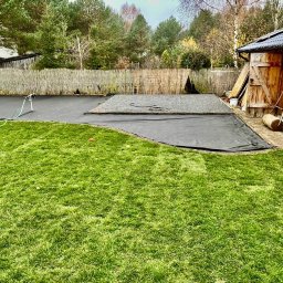 Volodymyr Stasyuk - Przygotowanie terenu pod budowę w ogrodzie: rozłożona czarna mata, fragment wysypany żwirem, trawnik i drewniana szopa w tle. Widok z poziomu trawy.