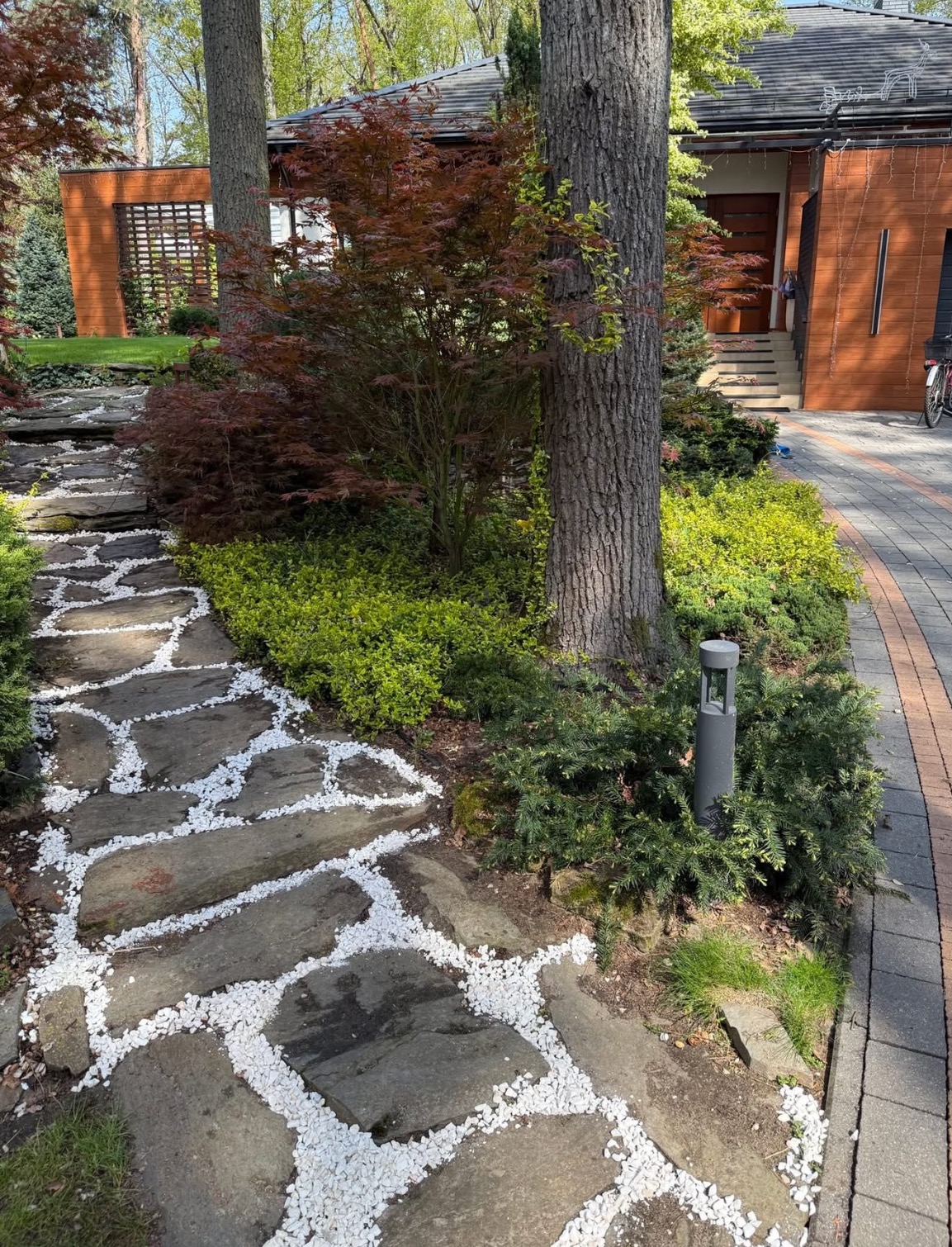 Kamienna ścieżka z białym żwirem w ogrodzie, prowadząca przez rabaty z krzewami i drzewami, oświetlona lampą ogrodową. W tle budynek z drewnianą elewacją.