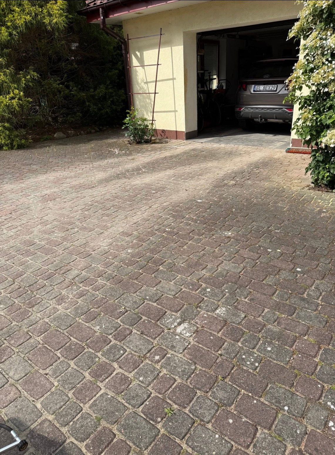 Ułożona kostka brukowa na podjeździe, z garażem w tle i roślinnością po bokach. Widoczne ślady użytkowania na kostce. Drabina oparta o ścianę garażu.