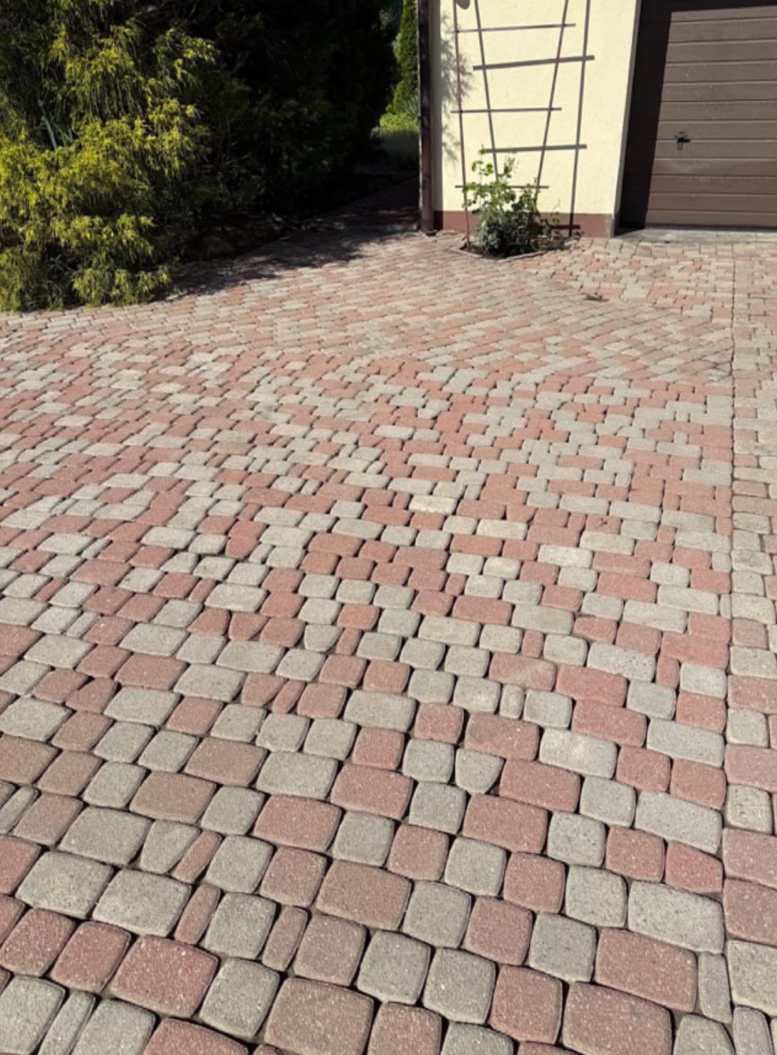 Ułożona kostka brukowa w odcieniach czerwieni i szarości tworzy wzór na podjeździe. Obok zielony krzew i fragment budynku z brązową bramą garażową i kratką na ścianie.