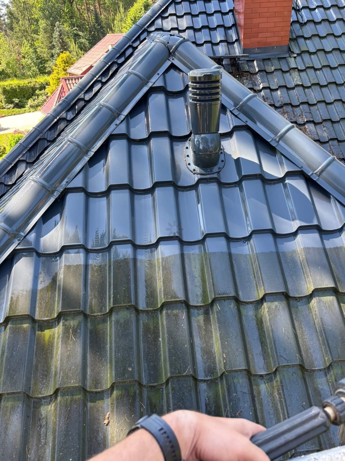 Częściowo umyta, ciemna dachówka z kominem wentylacyjnym. Górna część dachu jest czysta, dolna pokryta mchem. W tle widoczny dom i drzewa.