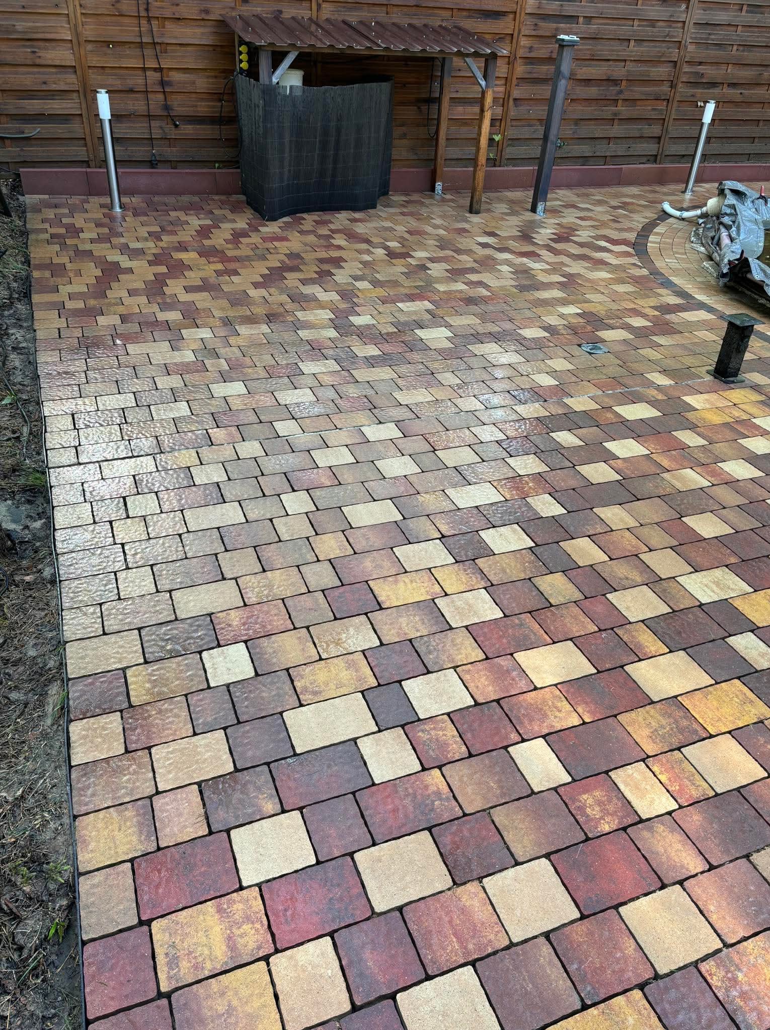 Kolorowa kostka brukowa ułożona na posesji, widoczne elementy oświetlenia i zadaszenie. Kostka w odcieniach brązu, beżu i czerwieni tworzy wzór na utwardzonej powierzchni.