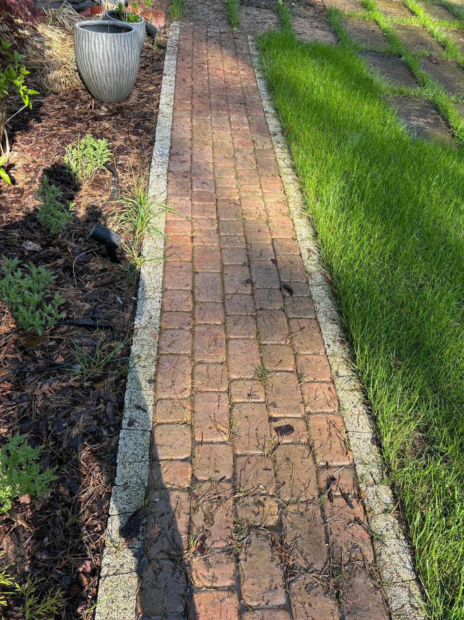 Ceglana ścieżka ogrodowa z obrzeżem, prowadząca przez trawnik. Obok rabata z korą, roślinami i donicami. Widoczne oświetlenie ogrodowe. Słoneczny dzień.