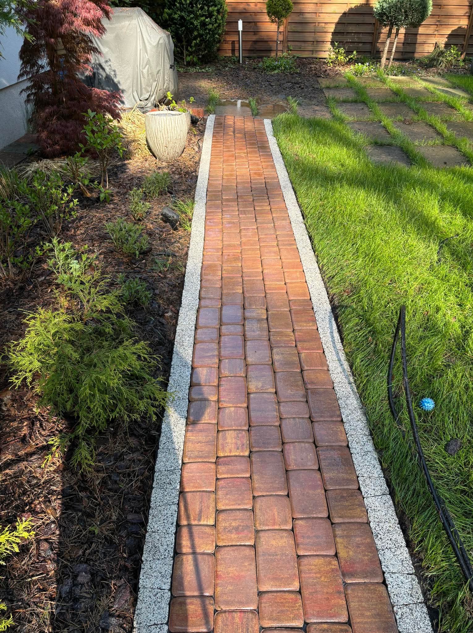 Ułożona ścieżka z kostki brukowej w ogrodzie, obrzeża z jasnego kruszywa. Widok z góry, w tle trawnik i roślinność.