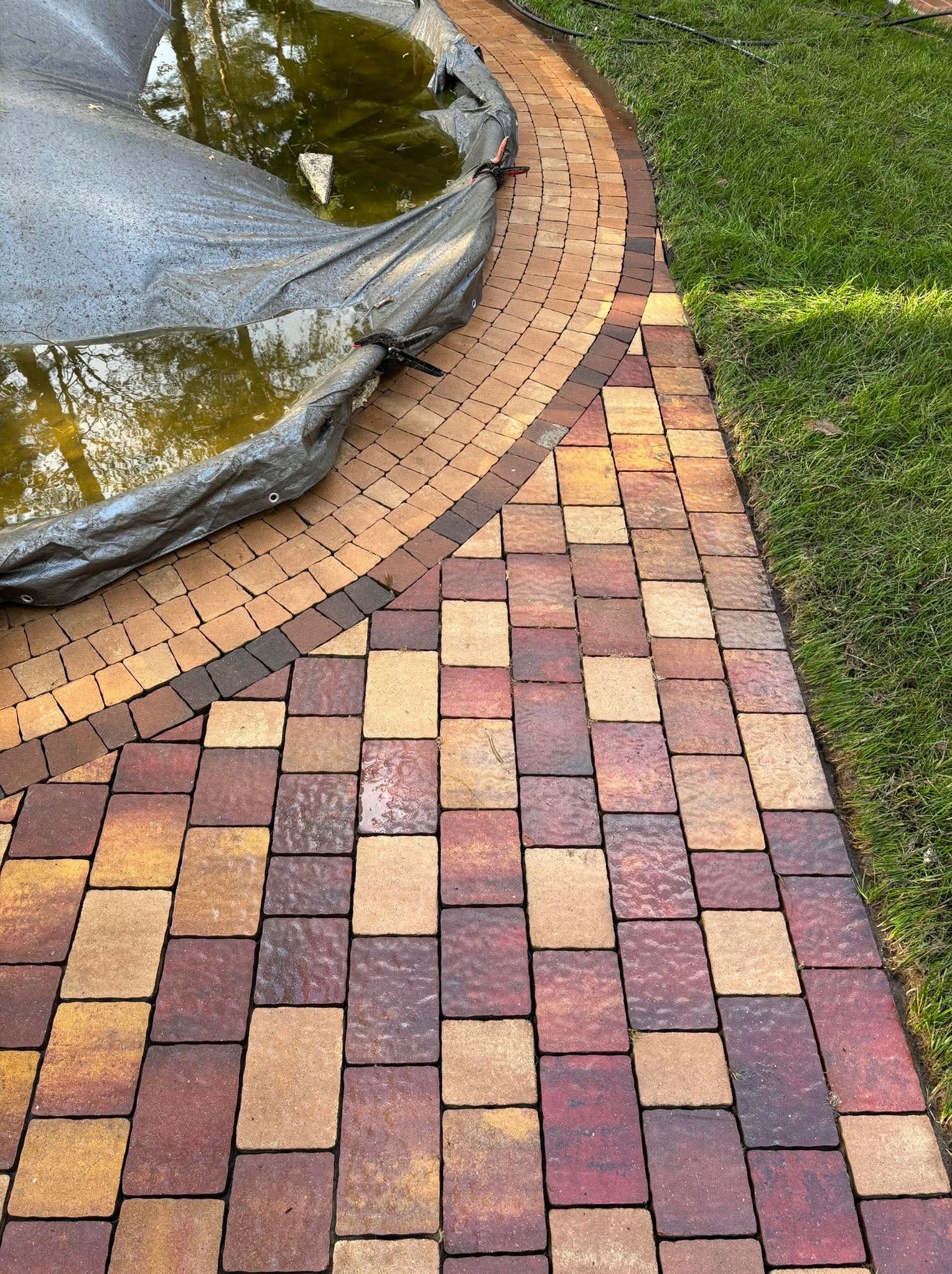 Kolorowa kostka brukowa tworzy ścieżkę obok trawnika i oczka wodnego z folią. Widok z góry, skupienie na wzorze i kolorystyce kostki.