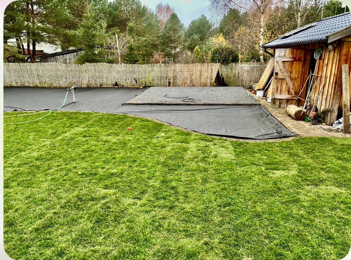 Przygotowanie terenu pod budowę w ogrodzie: rozłożona czarna mata, fragment wysypany żwirem, trawnik i drewniana szopa w tle. Widok z poziomu trawy.