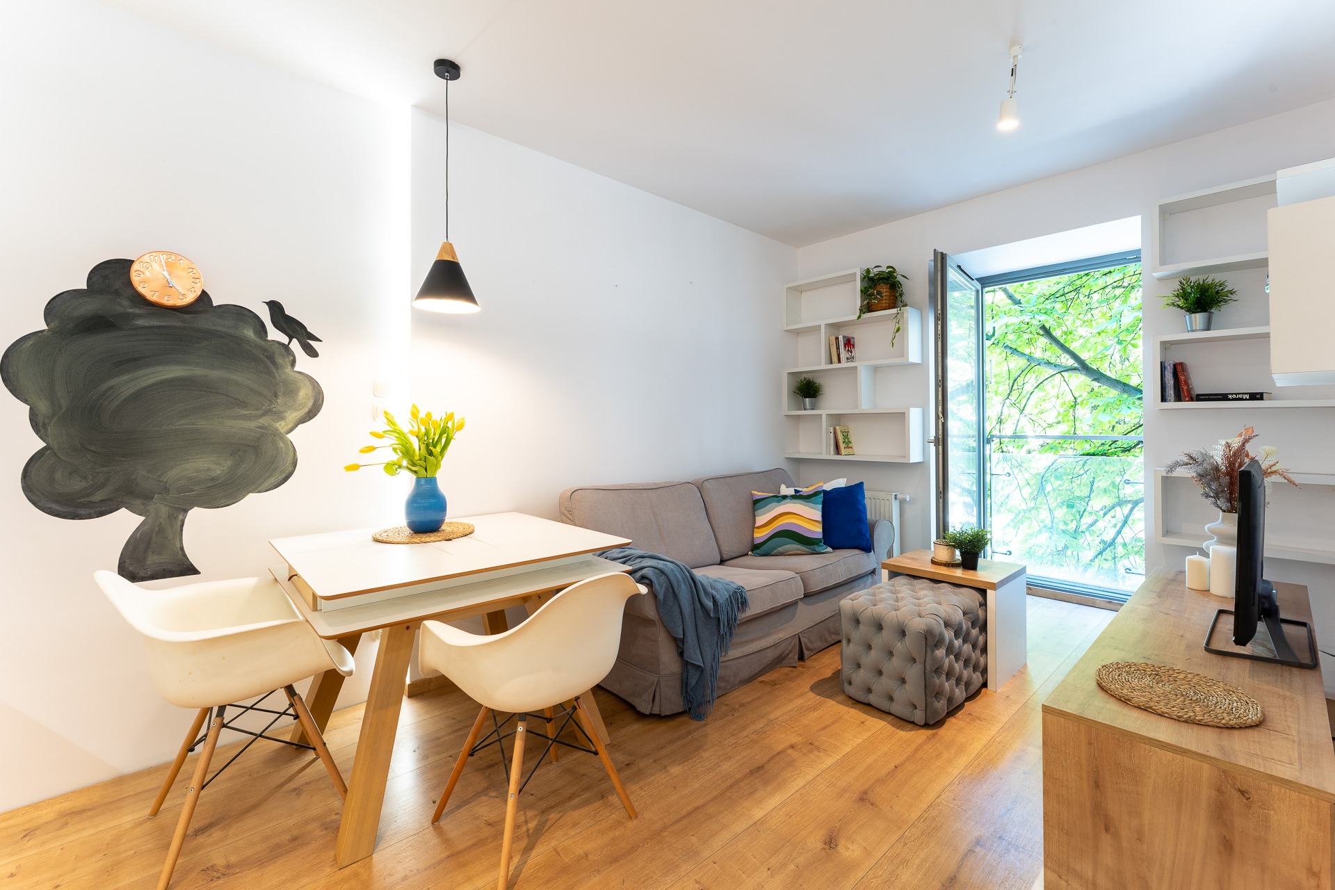 Jasne wnętrze mieszkania z sofą, stołem i dekoracją ściany w formie drzewa z zegarem. Widok na zieleń przez okno.