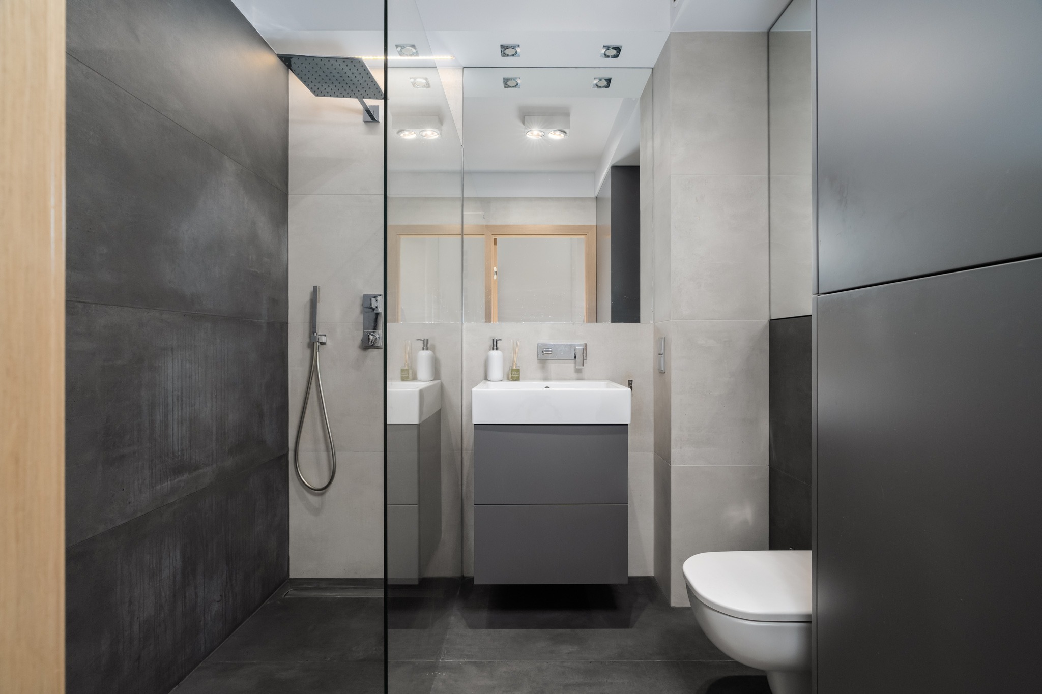 Nowoczesna łazienka z szarą armaturą, kabiną prysznicową typu walk-in i podwieszaną szafką z umywalką. Minimalistyczny design z akcentami betonu.