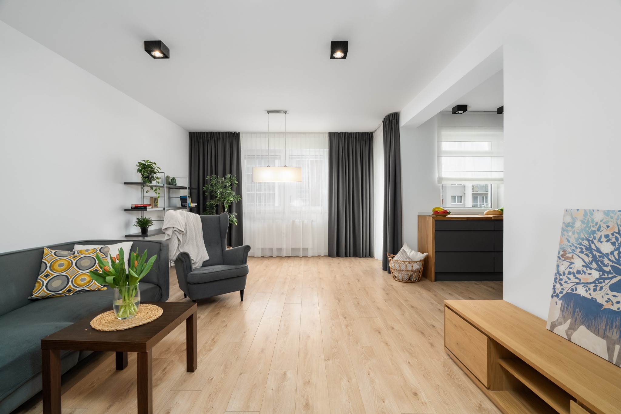 Jasne wnętrze z drewnianą podłogą, szarą sofą i fotelem, oświetlone światłem dziennym. Widoczne kwiaty w wazonie i dekoracje.