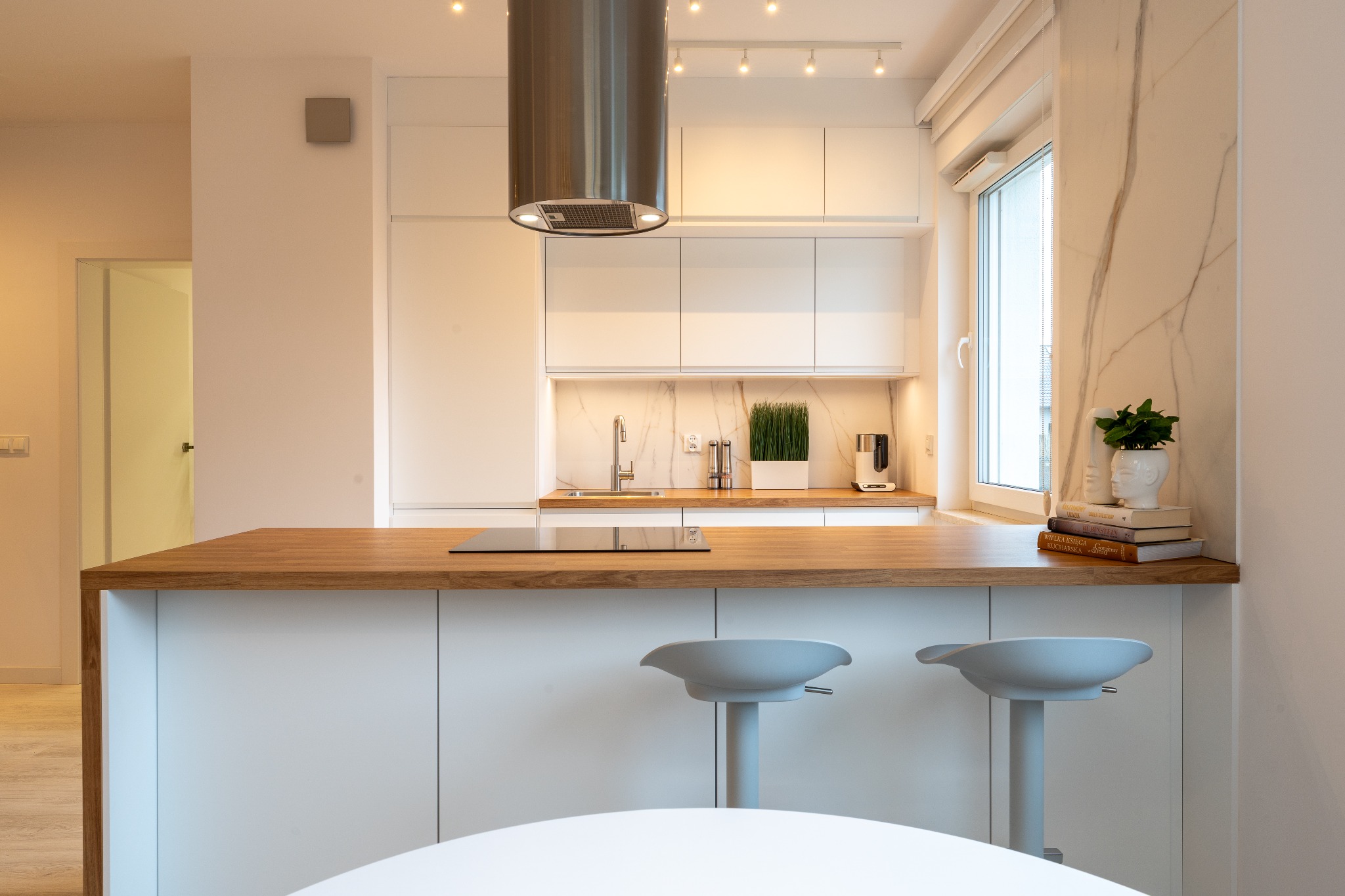 Jasna kuchnia z drewnianym blatem i marmurowym wykończeniem, nowoczesny design z hokerami i dekoracjami roślinnymi przy oknie. Minimalistyczny styl.