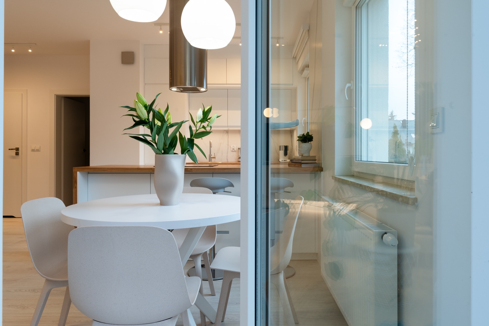 Jasne, nowoczesne wnętrze z okrągłym stołem i kwiatami w wazonie, widok przez szklane drzwi na minimalistyczną kuchnię z drewnianym blatem.