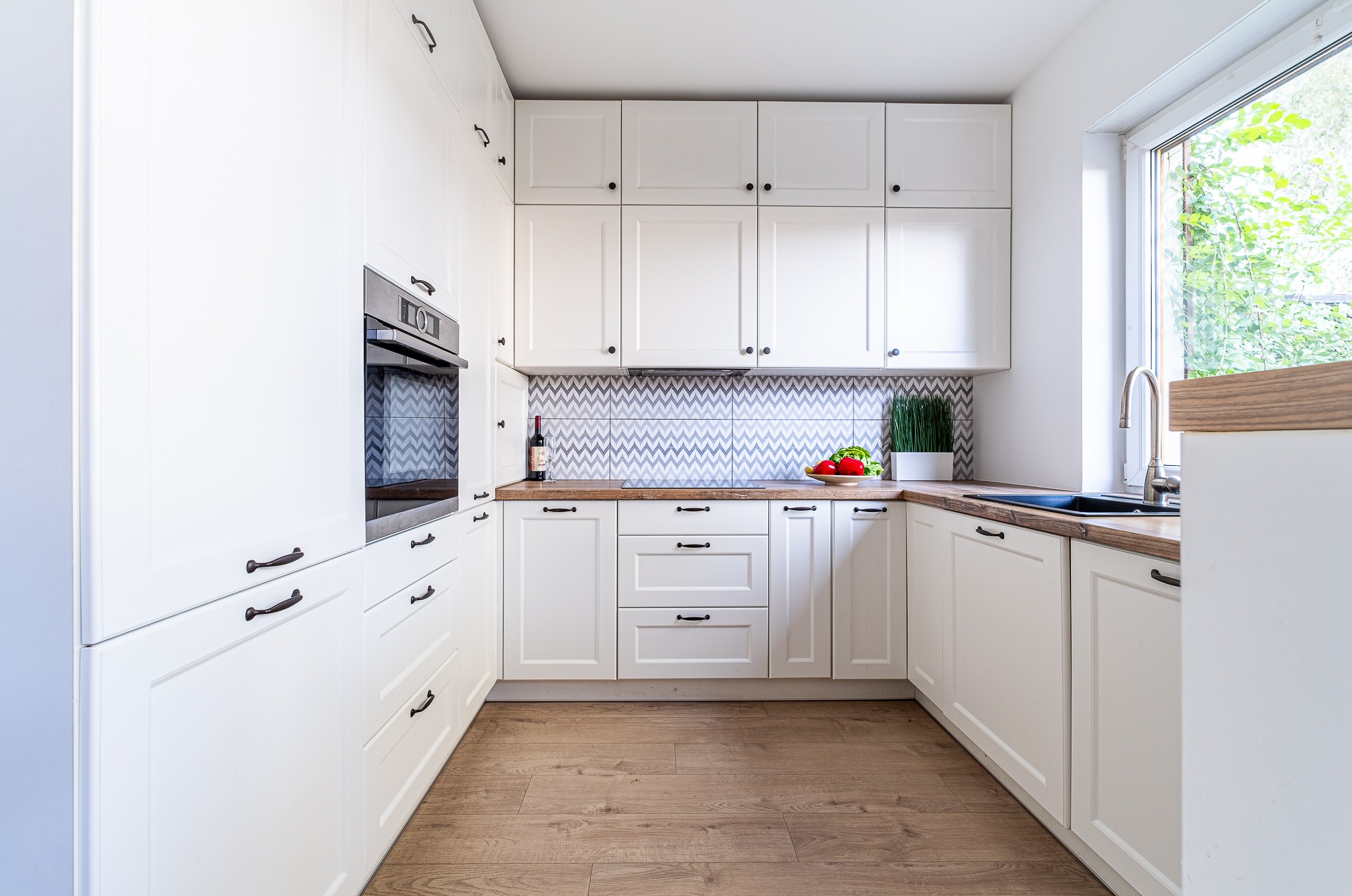Jasna kuchnia w stylu skandynawskim: białe szafki z ciemnymi uchwytami, drewniany blat, chevronowe płytki nad blatem, butelka wina, warzywa i zioła w doniczce.