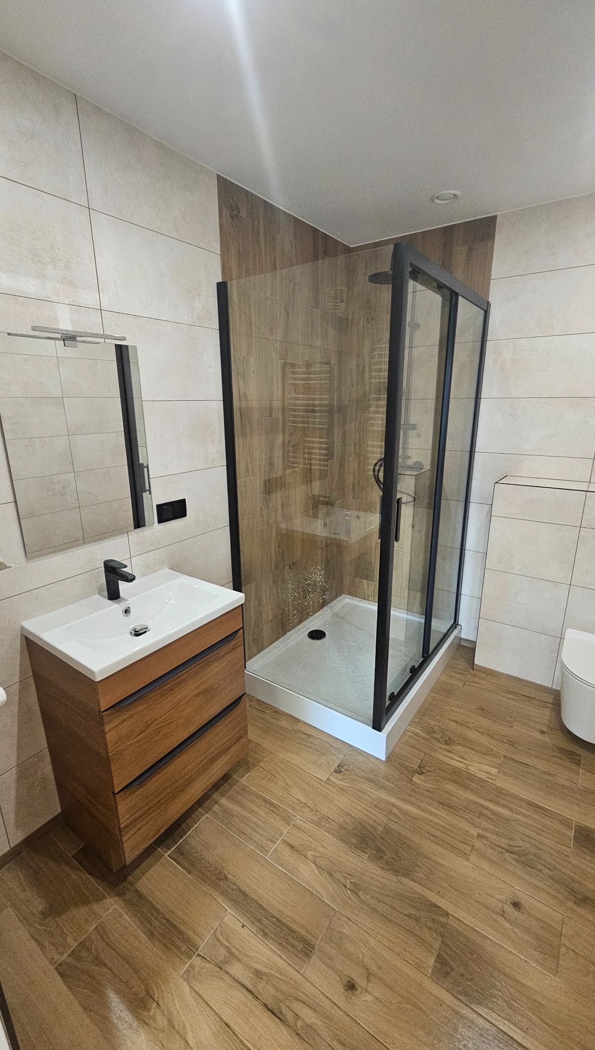 Nowoczesna łazienka z kabiną prysznicową typu walk-in, drewnopodobnymi płytkami na podłodze i ścianach oraz szafką z umywalką o minimalistycznym designie.