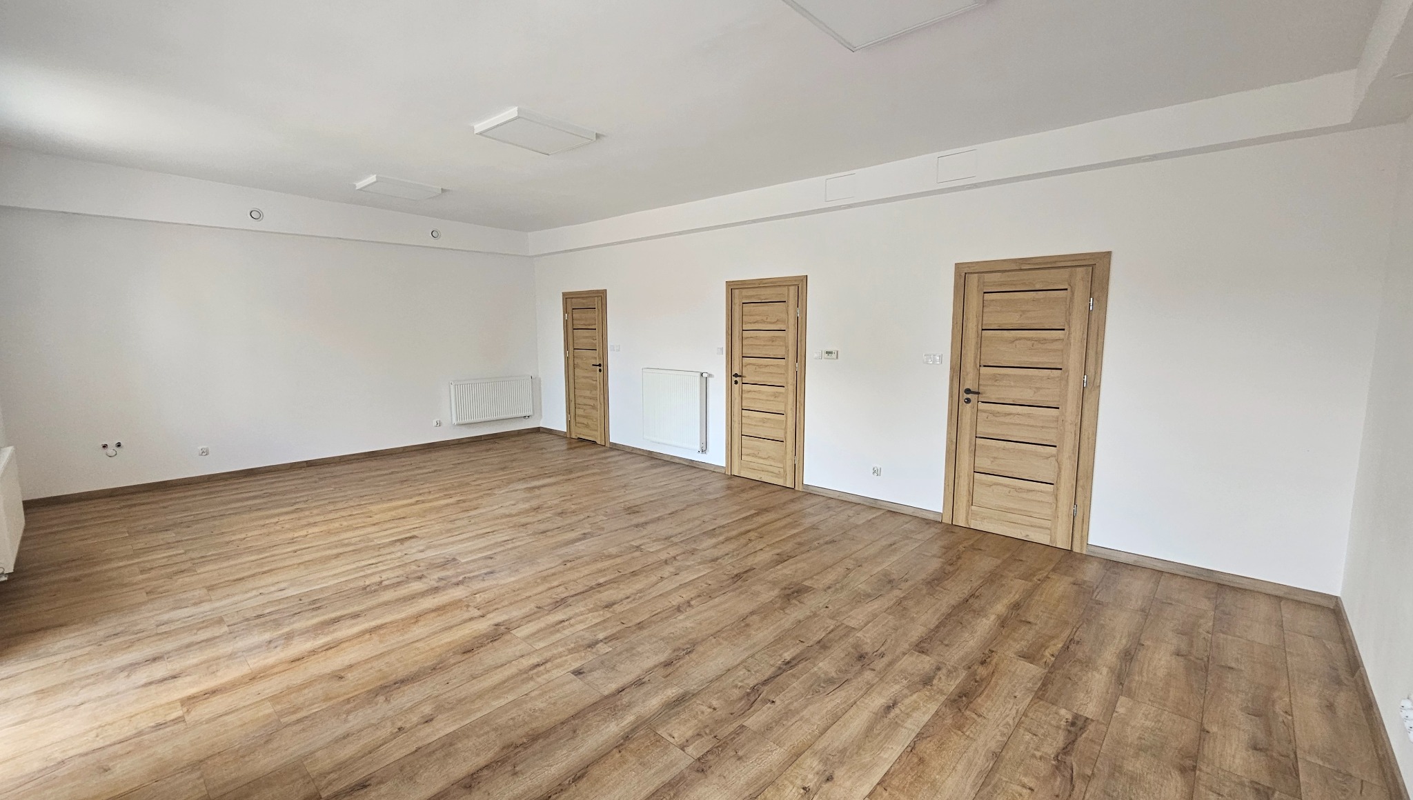 Jasne, przestronne wnętrze po remoncie: drewniana podłoga, białe ściany i trzy nowe drzwi z poziomymi panelami. Minimalistyczny design, widoczne grzejniki na ścianach.