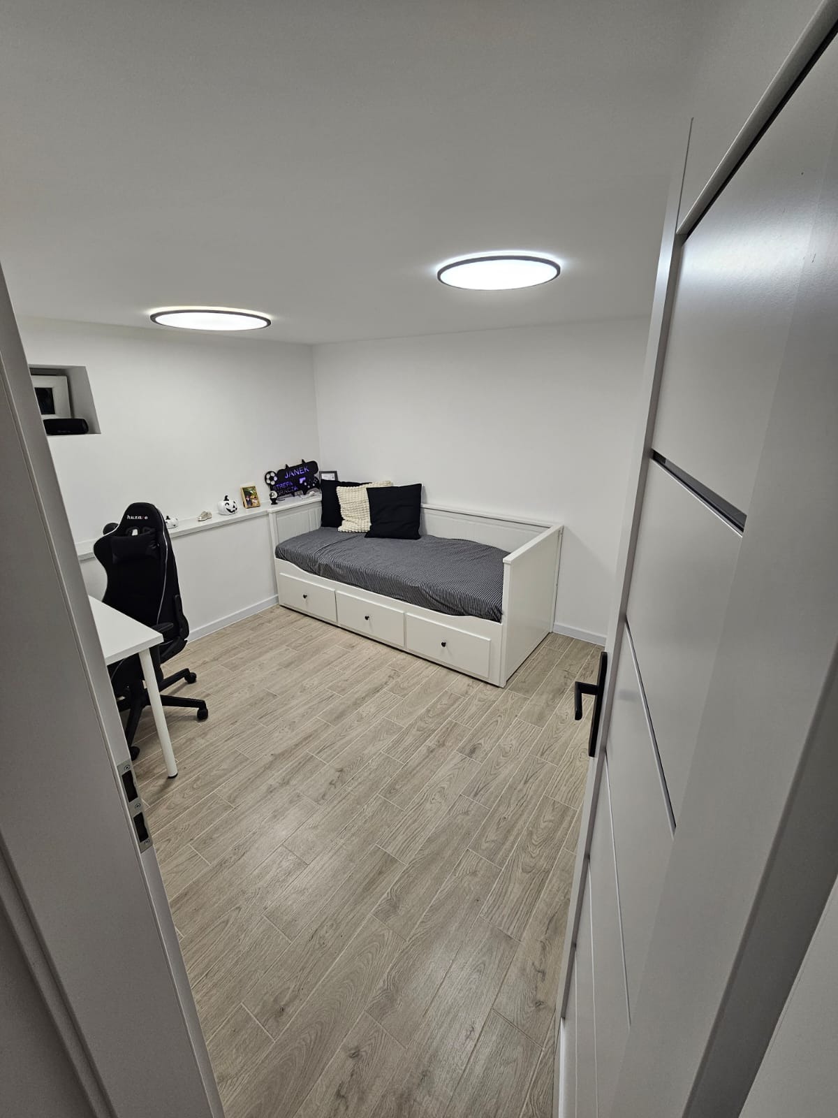 Pokój po remoncie: drewnopodobne panele, białe ściany, łóżko z szufladami i biurko z krzesłem gamingowym. Widok zza uchylonych, białych drzwi.