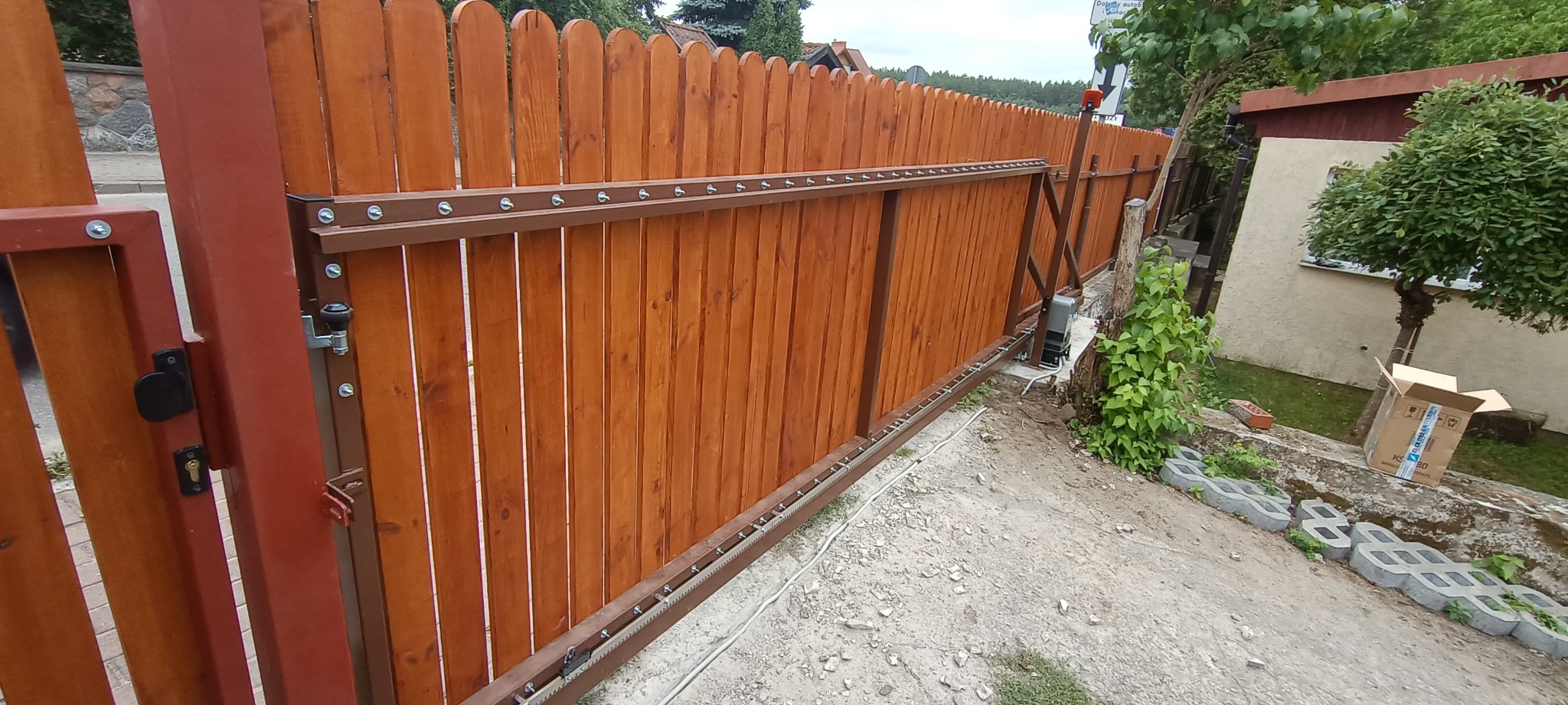 Brązowa, drewniana brama przesuwna z automatyką, widoczna z boku. Brama zamontowana na posesji, z widocznym mechanizmem i elementami mocującymi.