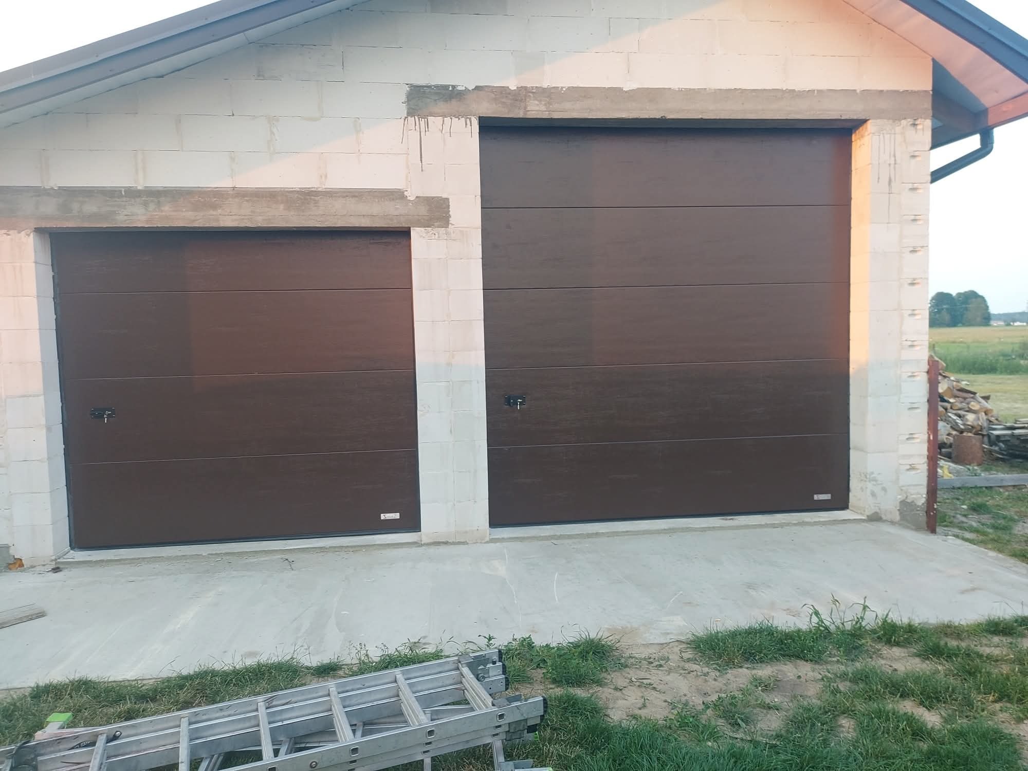 Dwie brązowe, segmentowe bramy garażowe w budynku z białych pustaków. Jedna brama szersza od drugiej. Na dole kadru drabina. Widok z zewnątrz.