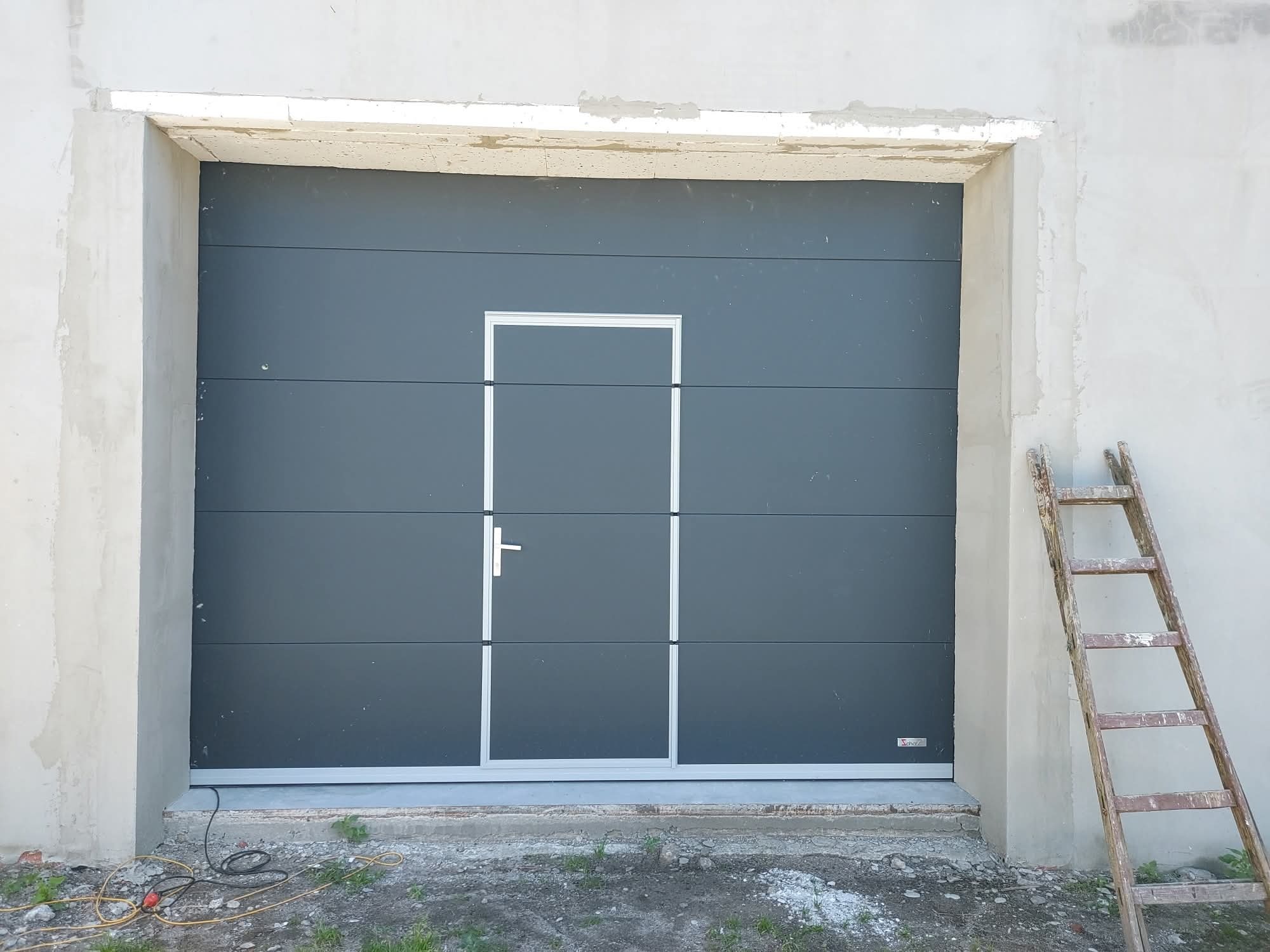 Szara brama garażowa segmentowa z drzwiami i klamką, widoczna drabina oparta o ścianę, surowy stan wykończenia budynku, kabel elektryczny na ziemi.