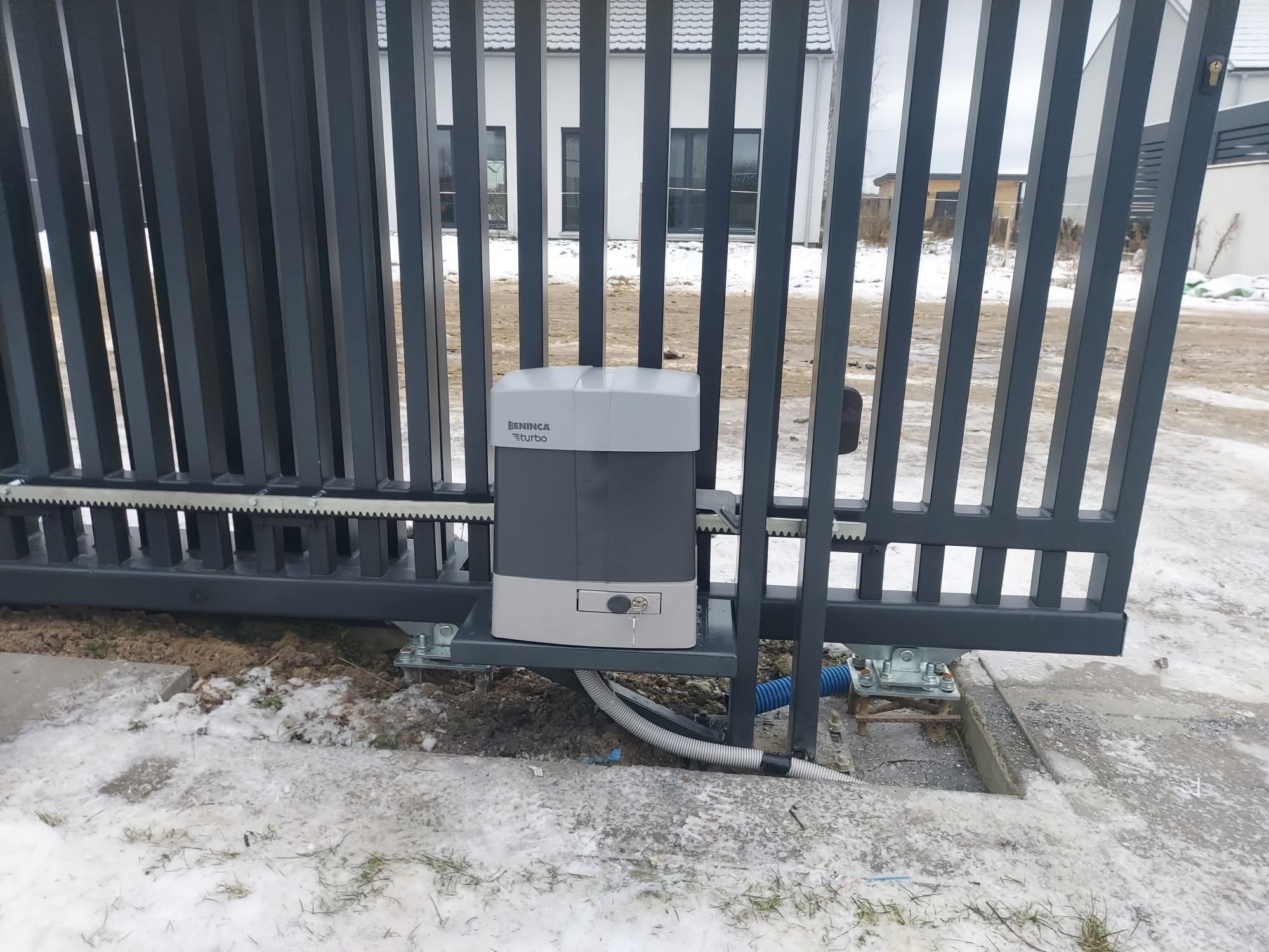 Szara, przesuwna brama z napędem Beninca Turbo. Widoczny mechanizm i klucz. W tle dom z szarą elewacją. Zima, śnieg na ziemi.