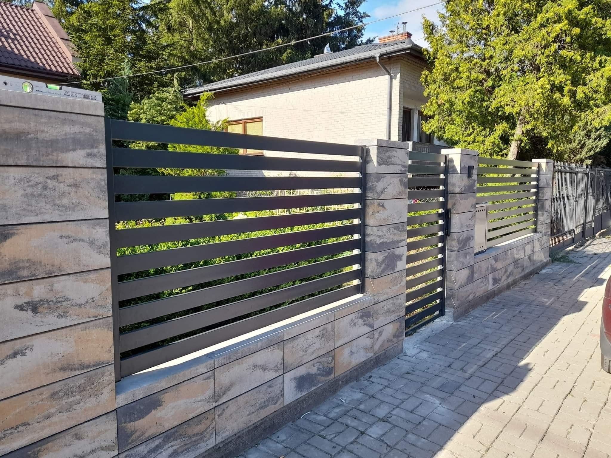 Nowoczesne ogrodzenie panelowe z poziomymi, grafitowymi przęsłami, osadzone na słupkach z jasnoszarej kostki. Widoczny fragment posesji i kostka brukowa.