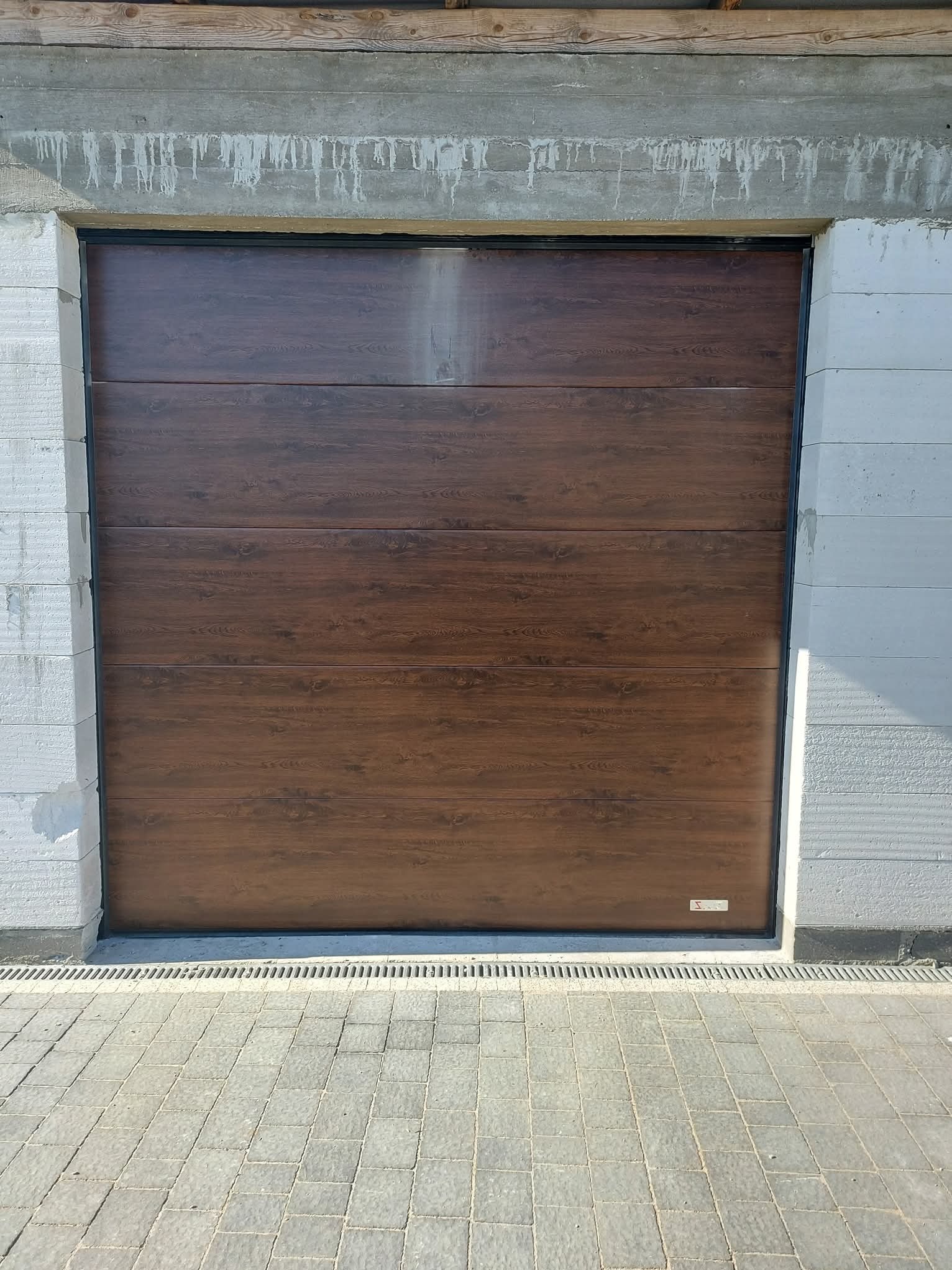 Zamknięta, segmentowa brama garażowa w kolorze ciemnego drewna, zamontowana w surowym otworze budynku z betonu komórkowego. Widoczna kostka brukowa przed bramą.