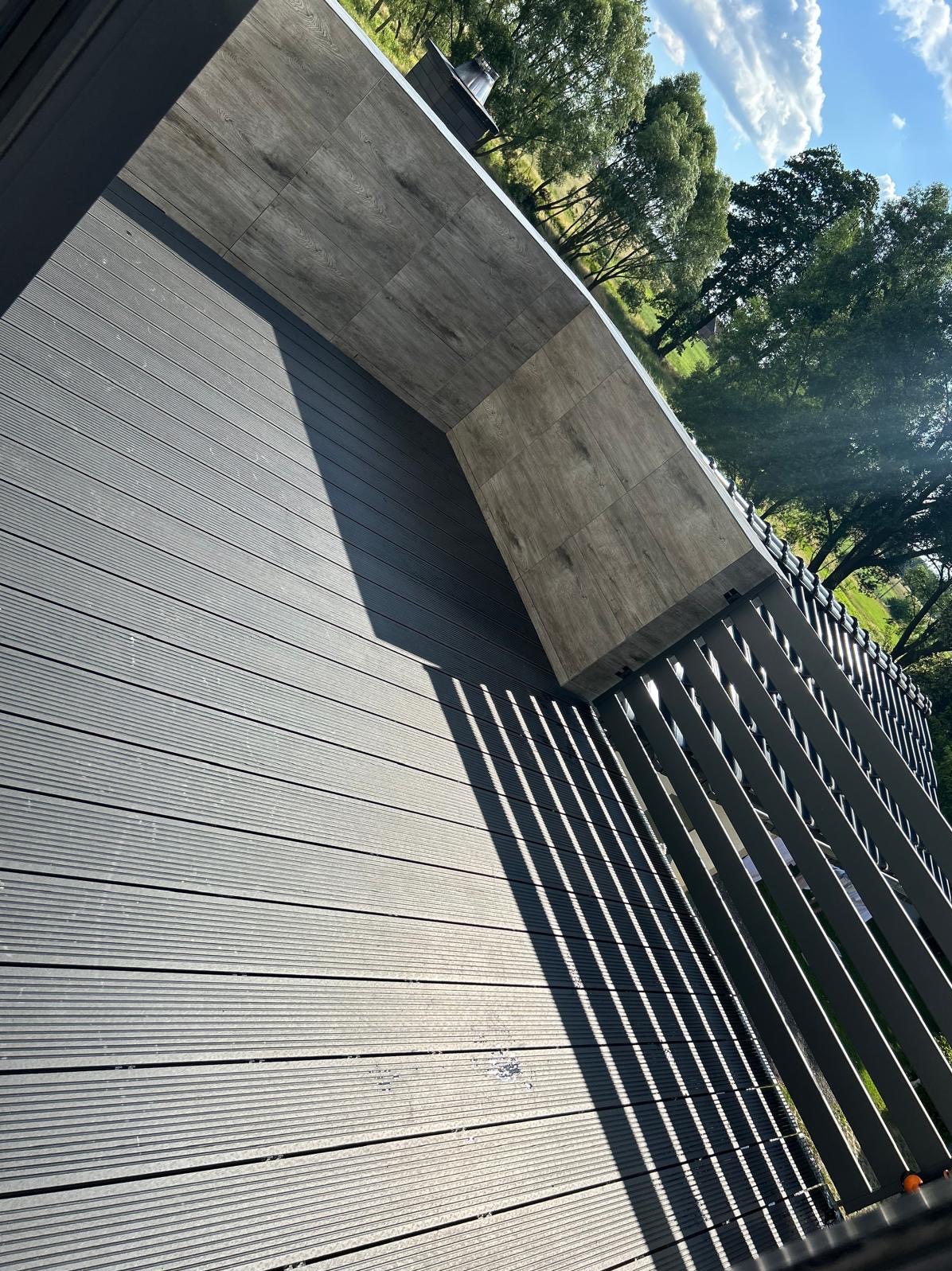 Nowoczesny balkon z szarymi deskami kompozytowymi i płytkami imitującymi drewno na ścianie, z metalową balustradą i widokiem na zieleń w tle.