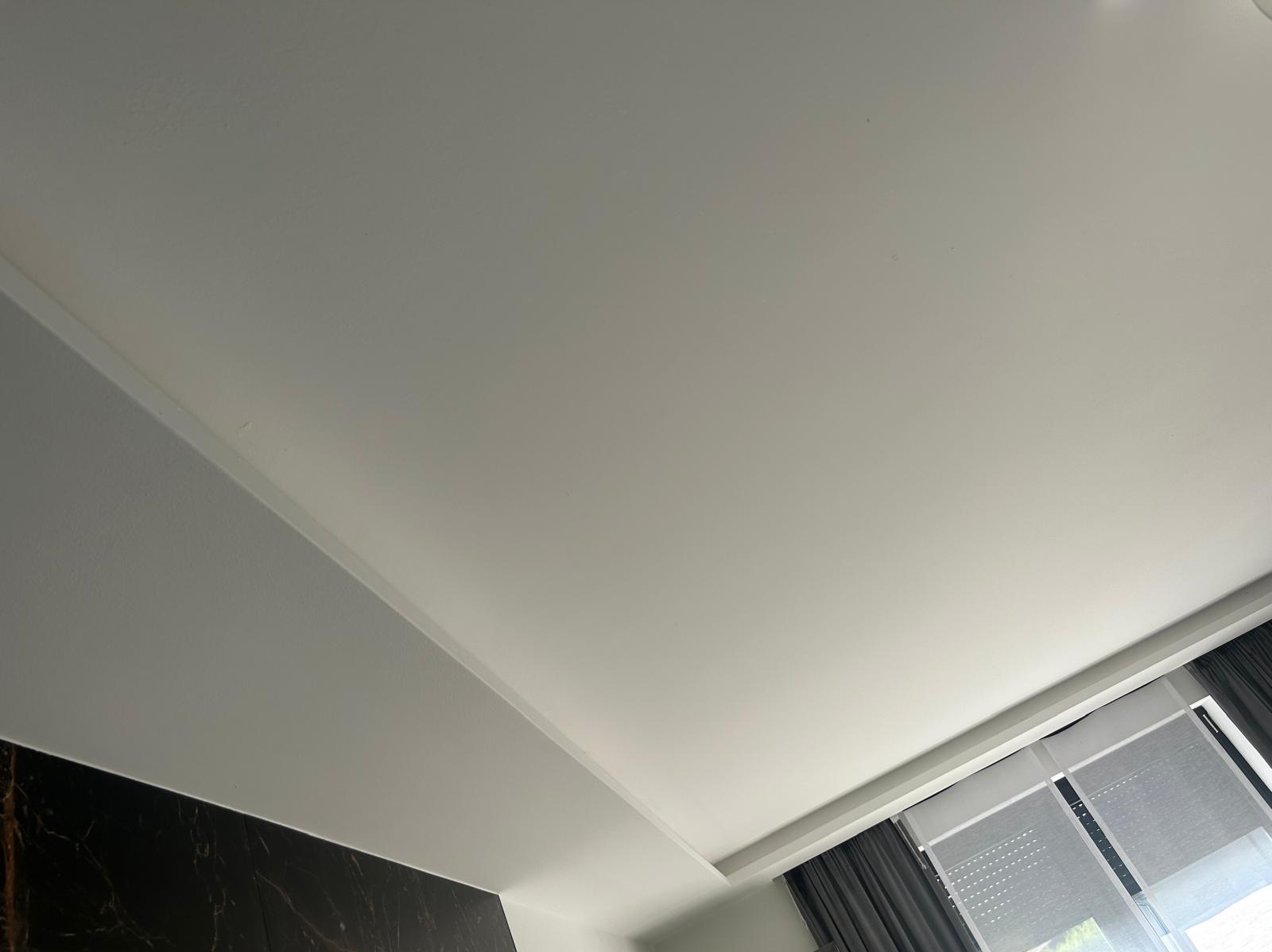 Biały sufit z listwami dekoracyjnymi i fragmentem okna z zasłonami, widziany pod kątem. Minimalistyczny design wnętrza z elementami wykończeniowymi.