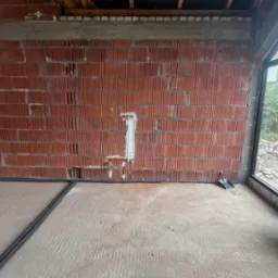 Wewnętrzna ściana z cegły z pionowo zamontowaną puszką instalacyjną i pomarańczowymi przewodami. Czarne kable przy podłodze. Okno z widokiem na zieleń.