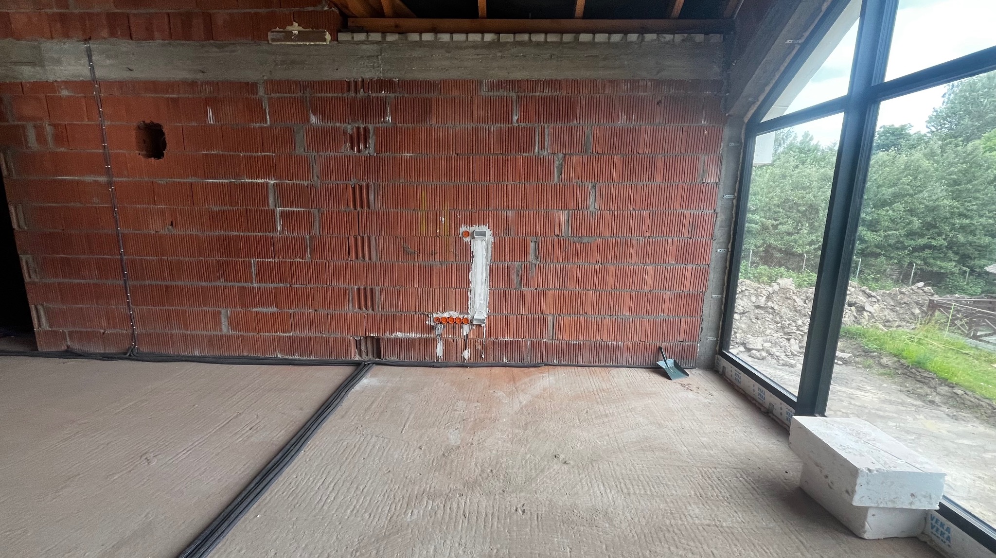 Wewnętrzna ściana z cegły z pionowo zamontowaną puszką instalacyjną i pomarańczowymi przewodami. Czarne kable przy podłodze. Okno z widokiem na zieleń.