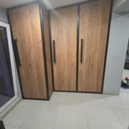 Trzydrzwiowa szafa z drewnopodobnymi frontami i czarnymi obramowaniami, widoczna w rogu pomieszczenia z jasną podłogą.