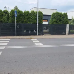 Nowoczesna, pozioma brama wjazdowa z metalowymi lamelami w kolorze antracytowym, widziana z perspektywy ulicy z przejściem dla pieszych i zielonymi tujami w tle.