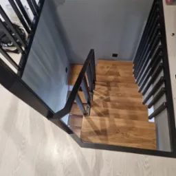 Drewniane schody z czarną, metalową balustradą, ujęte z góry. Widoczne schody prowadzą w dół, oświetlone światłem naturalnym. Minimalistyczny design.