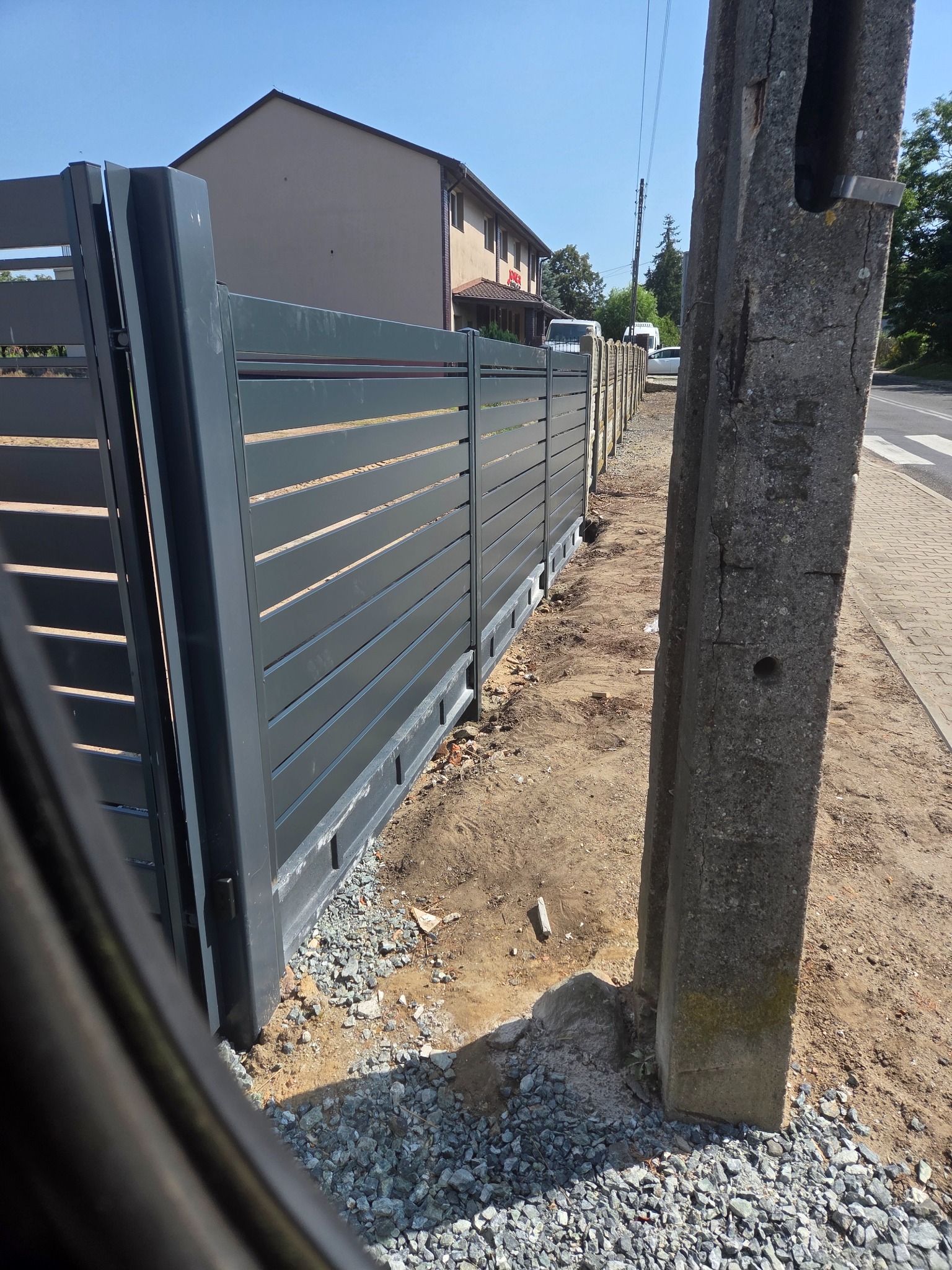 Nowoczesna brama wjazdowa z poziomych paneli w kolorze grafitowym, zamontowana przy posesji. Widoczny fragment ogrodzenia i słup energetyczny.